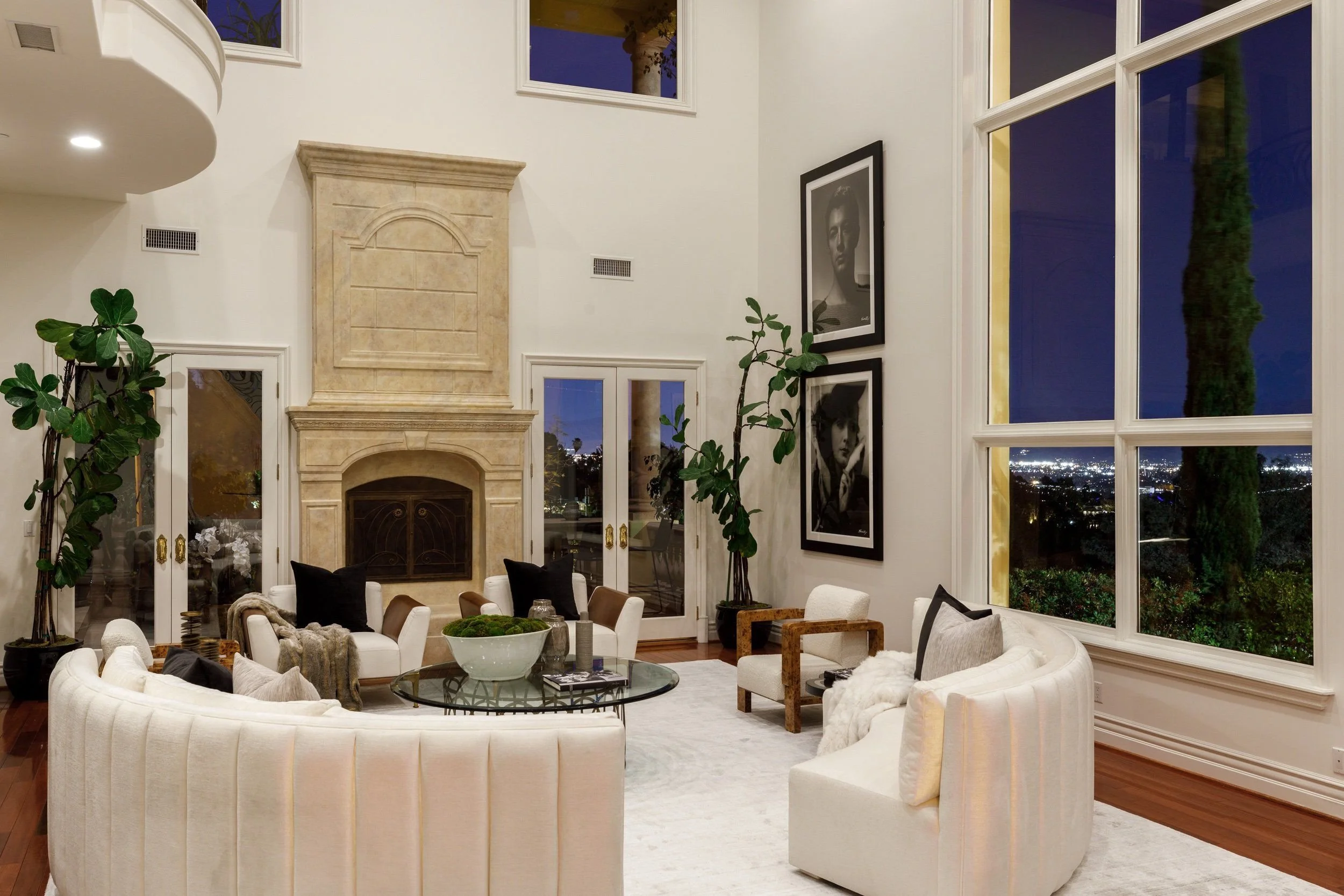 A spacious living room with a beige stone fireplace, large windows showing a nighttime cityscape, white upholstered sofas and armchairs with black and beige pillows, a glass coffee table, and framed black and white portraits on the wall. Green plants