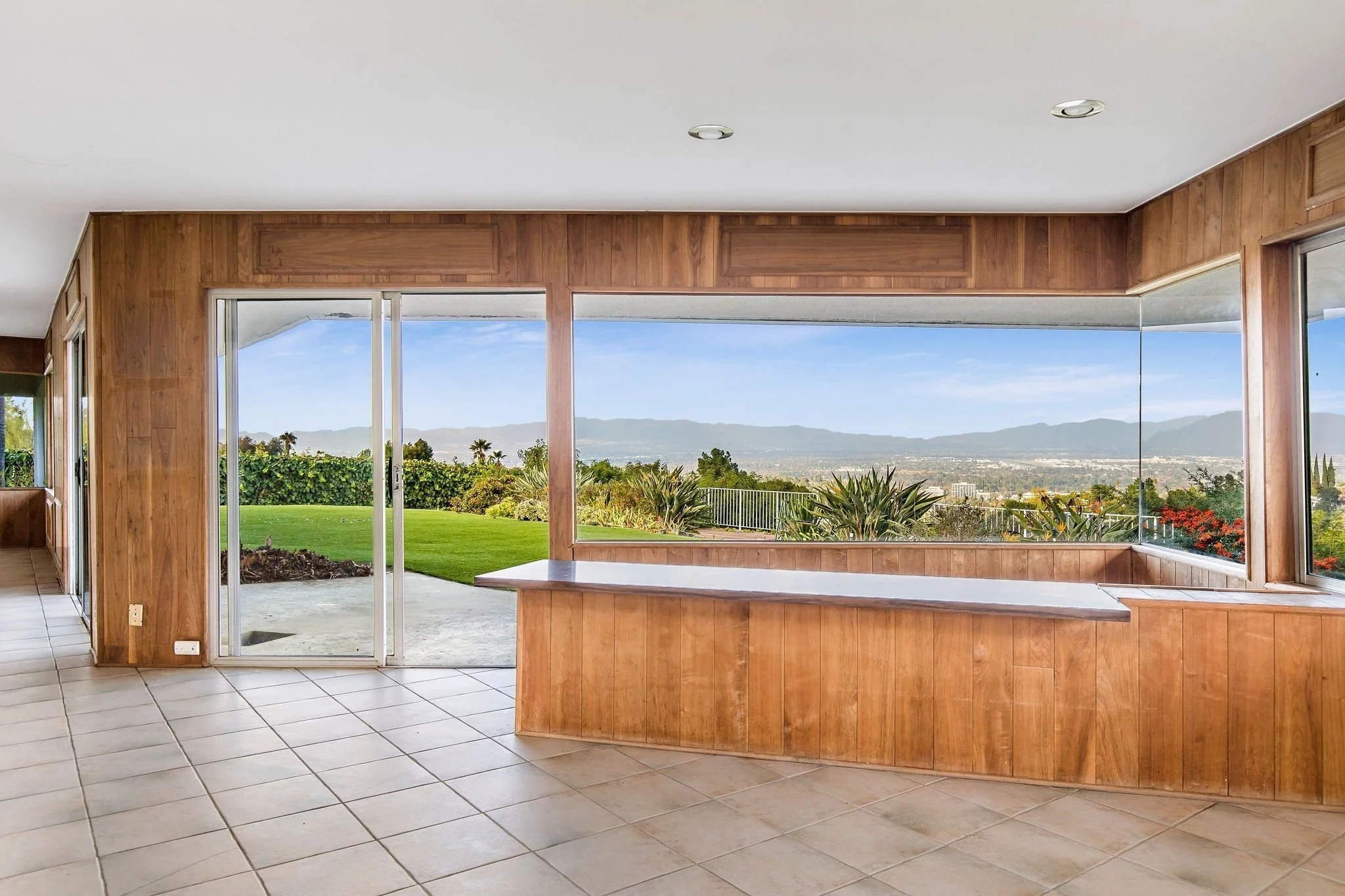 Room with large sliding glass doors showing a lush green backyard with plants and mountains in the distance, and a white ceiling with two recessed lights.