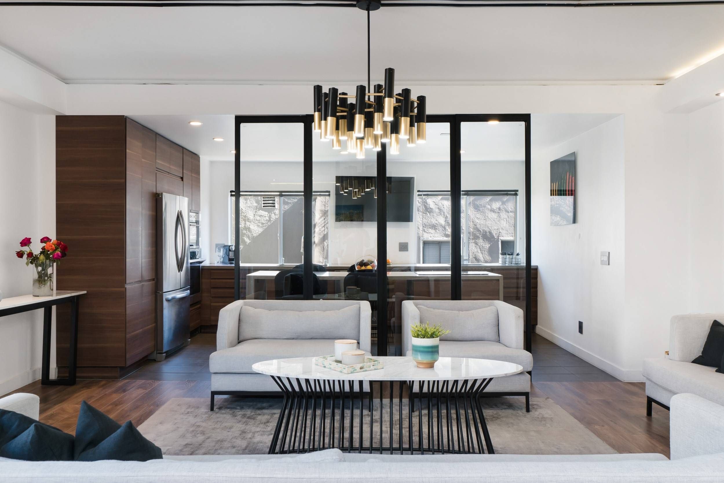 Modern living room with white sofa, black metal coffee table, and decorative pillows, adjacent to a dining area with a round table and white chairs. The background features a kitchen with wooden cabinets, a stainless steel refrigerator, and large win