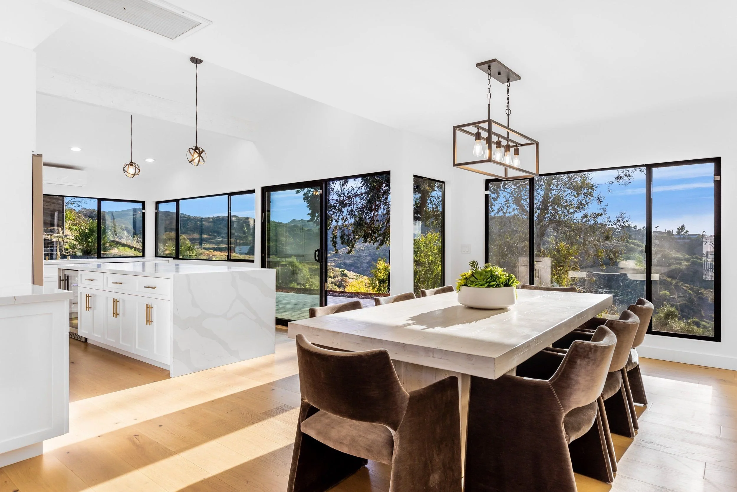 Bright, modern kitchen and dining area with large windows showing a scenic hill and trees outside. The kitchen has white cabinets and a white island. The dining table is light-colored, surrounded by beige upholstered chairs. Modern pendant and chande