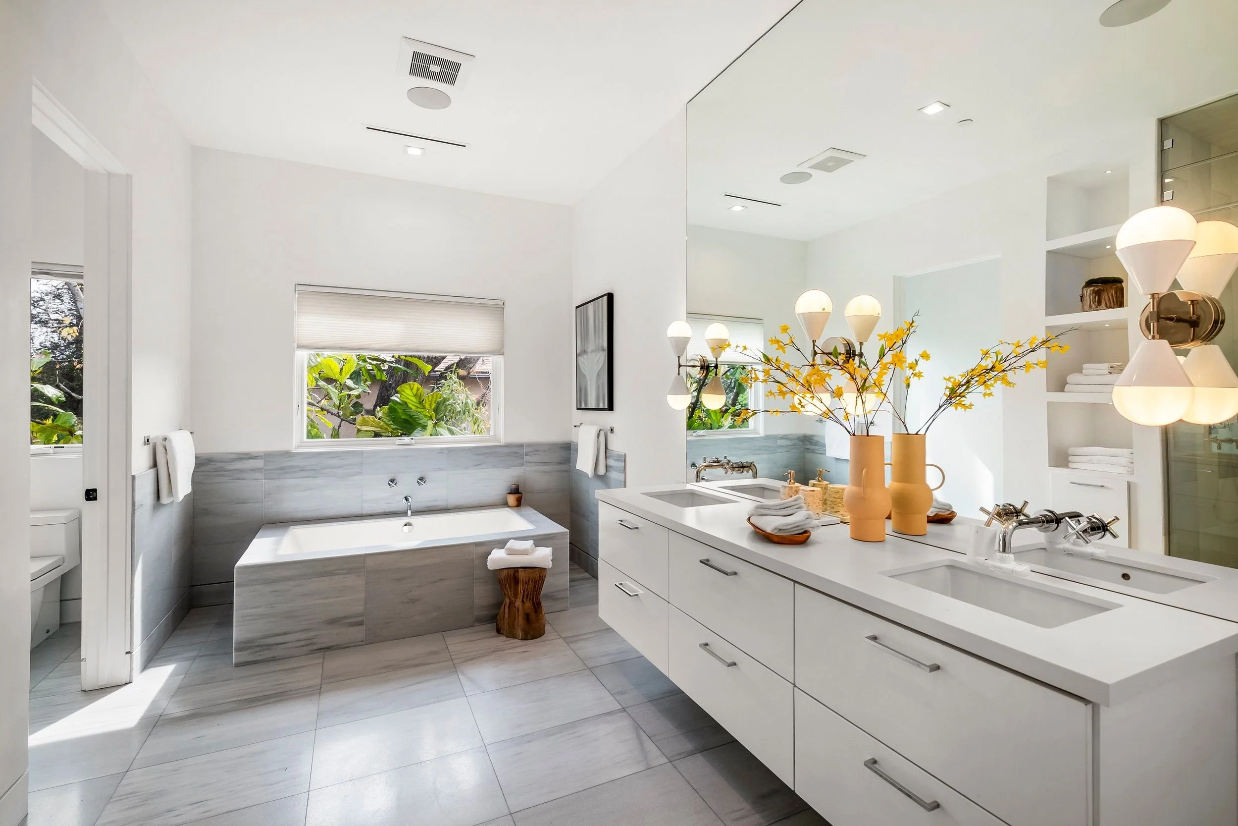 Modern bathroom with bathtub under window, white cabinetry, yellow vases with flowers, and a large mirror.