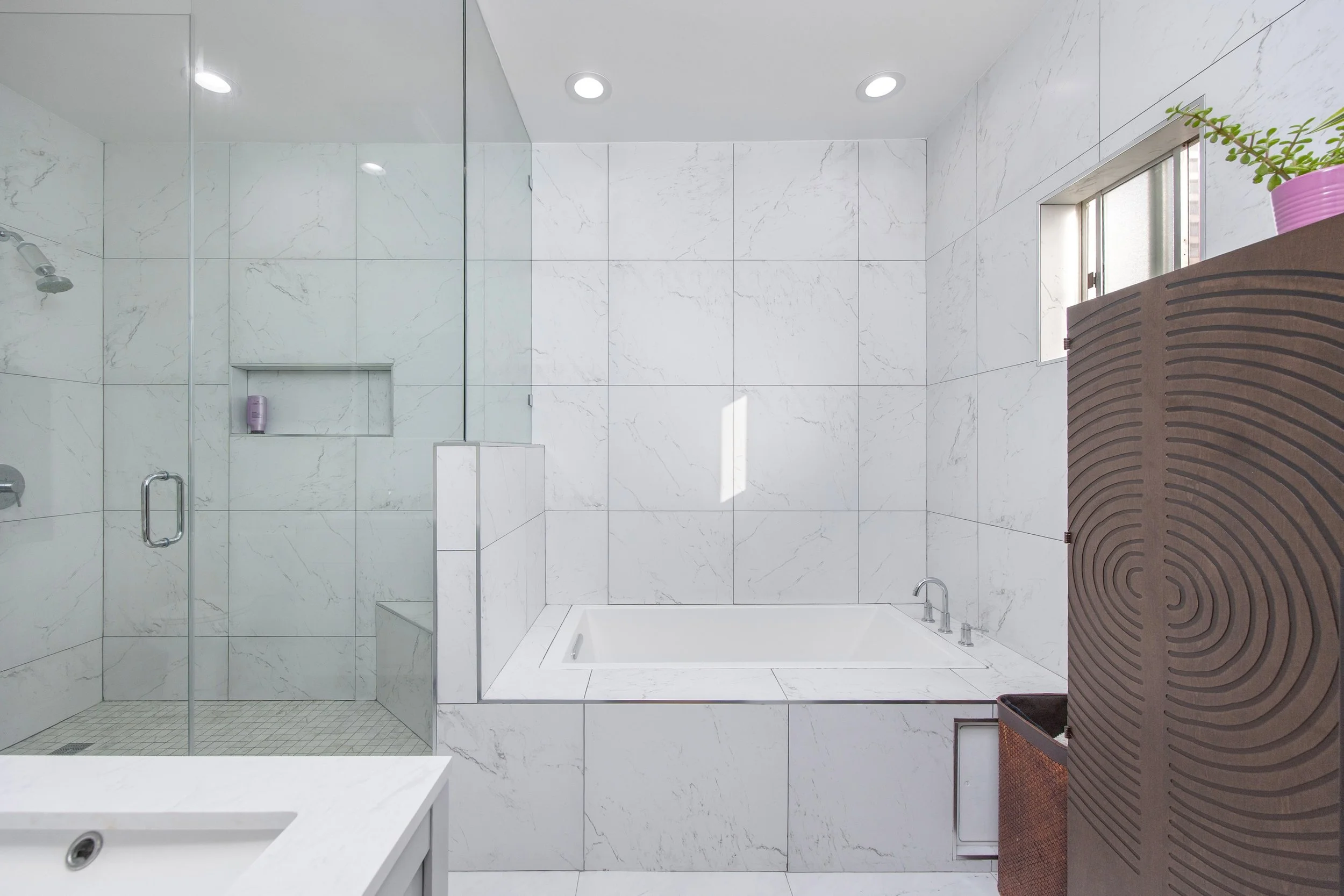 A modern bathroom with white marble walls and floor, a large bathtub, a glass-enclosed shower with a built-in shelf, a window with frosted glass, and a brown wicker basket with a plant in a pink pot on top.