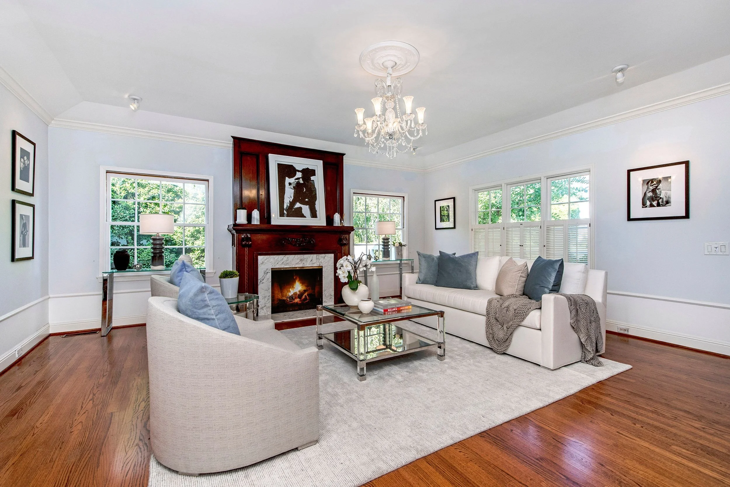 Living room with white sofa, gray pillows, glass coffee table, wooden floor, fireplace with artwork, and wall art, bright with large windows and a chandelier.