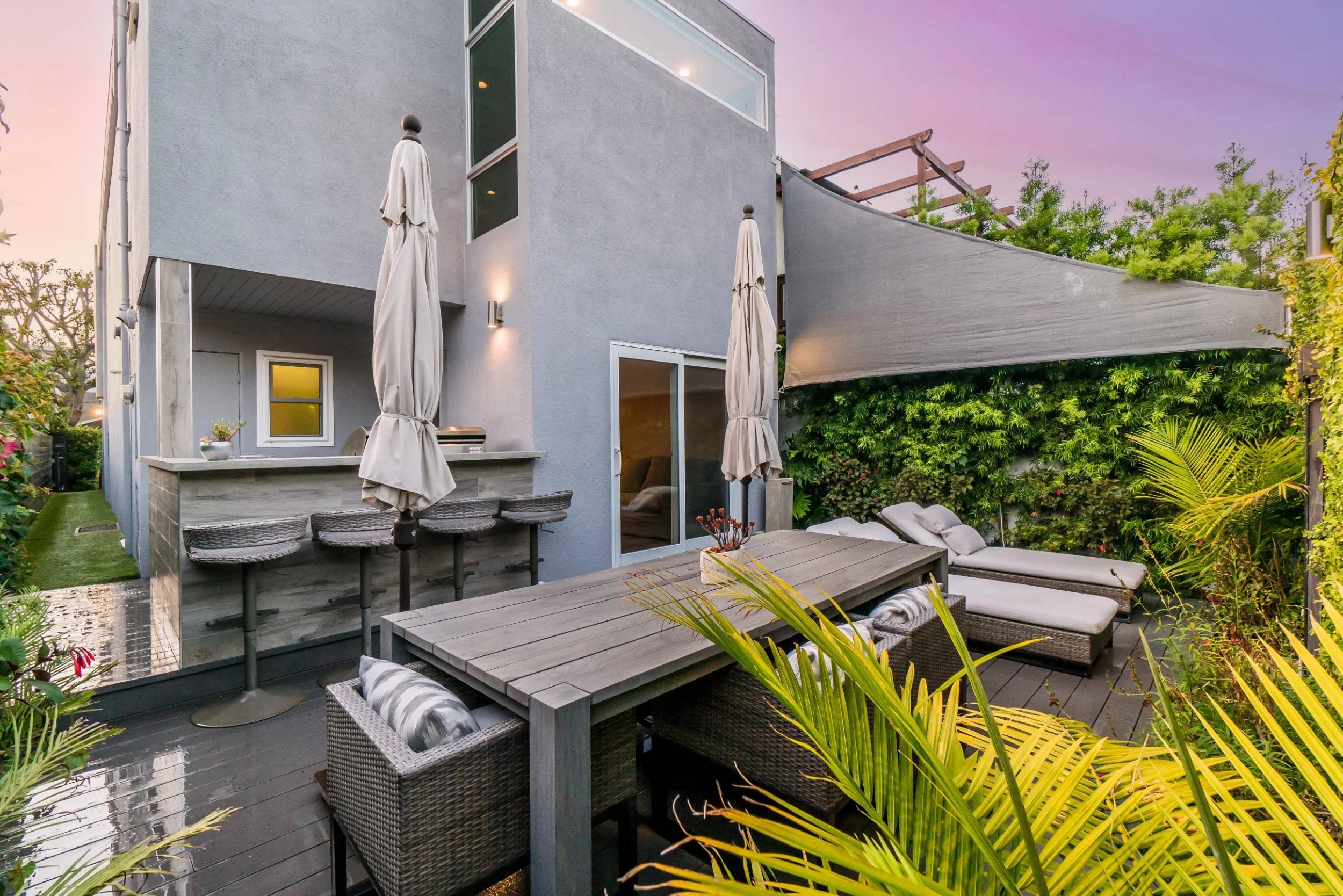A modern outdoor patio with a gray house in the background, featuring a bar area with barstools, a dining table with chairs, and lounge chairs, surrounded by lush greenery and purple sunset sky.