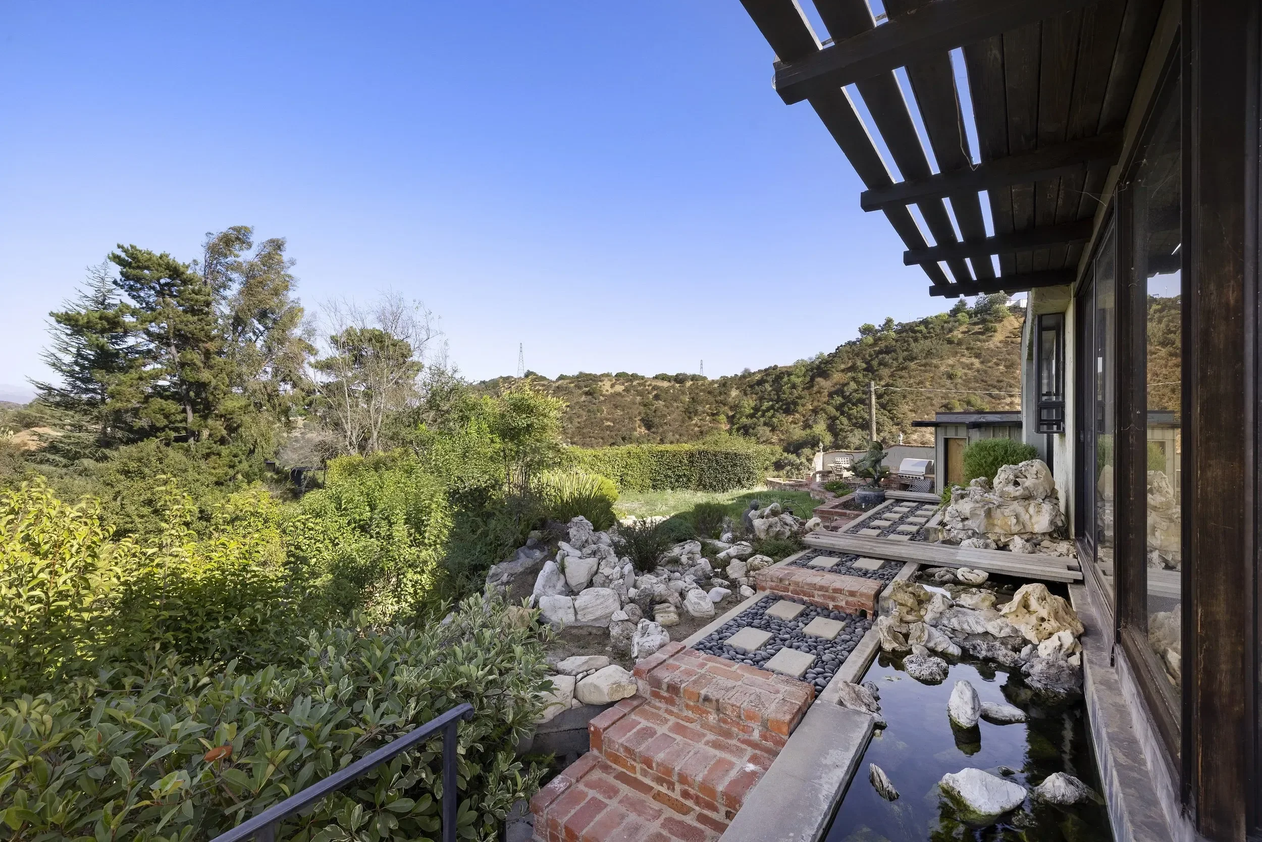 A backyard with a landscaped garden, a small pond with rocks, and a modern house with large windows and a wooden exterior. There are trees and hills in the background under a clear blue sky.