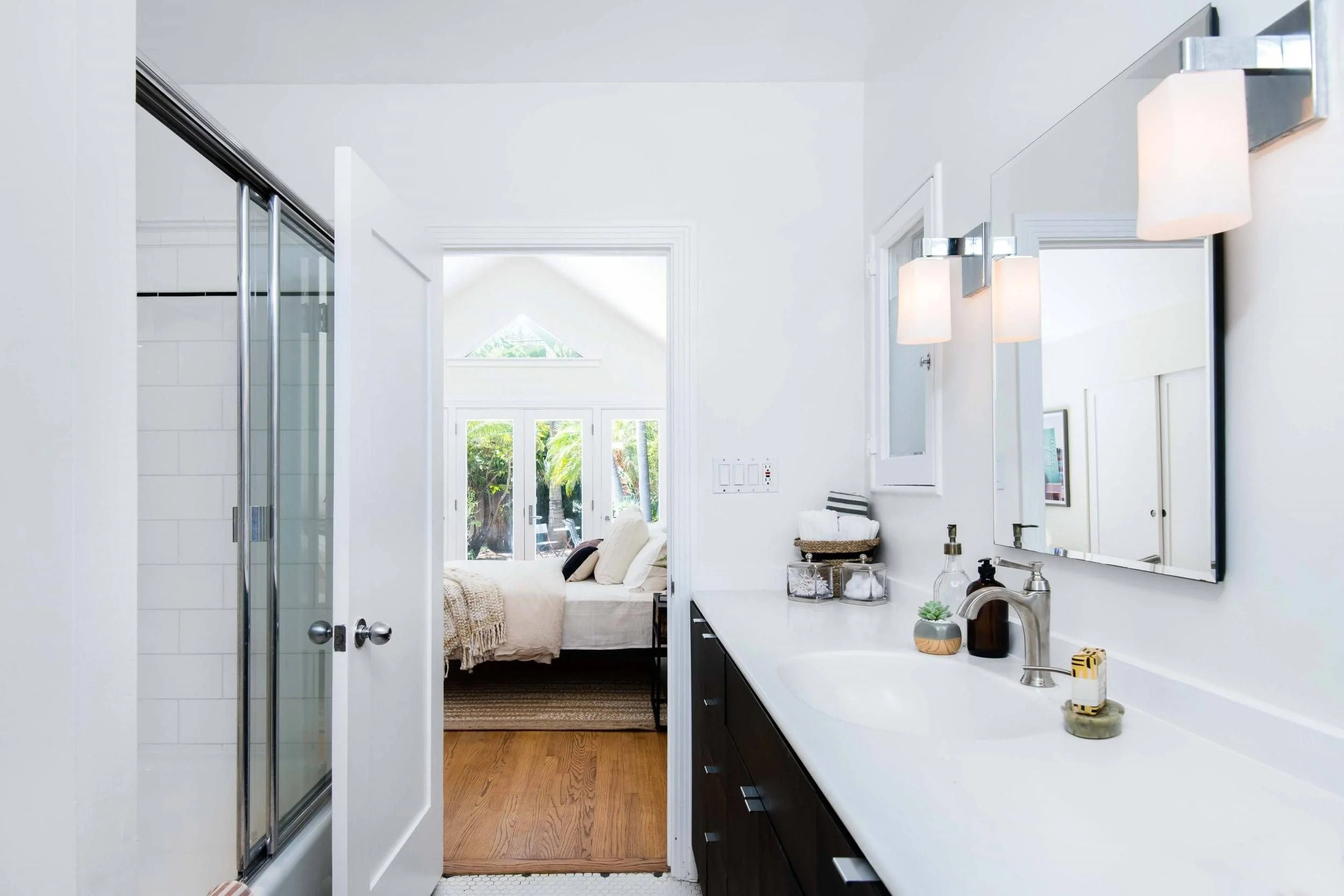 Modern bathroom with white walls, a large mirror, and black cabinets, opening to a bedroom with a bed, window, and outdoor view.