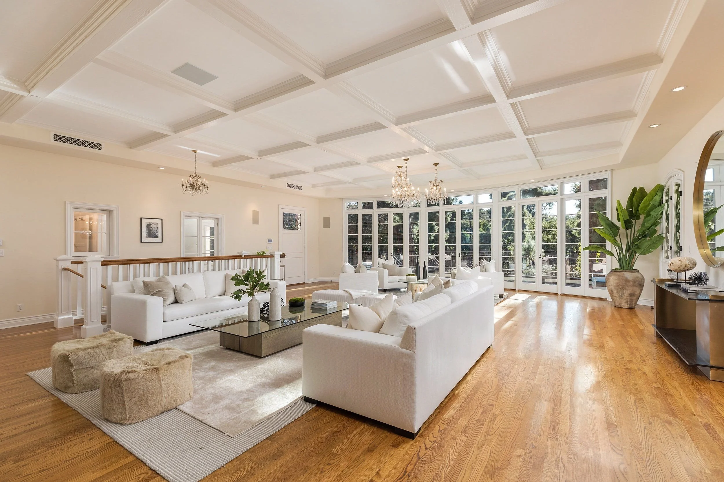 Bright spacious living room with white sofas, a glass coffee table, large windows, chandelier lighting, and wooden flooring.