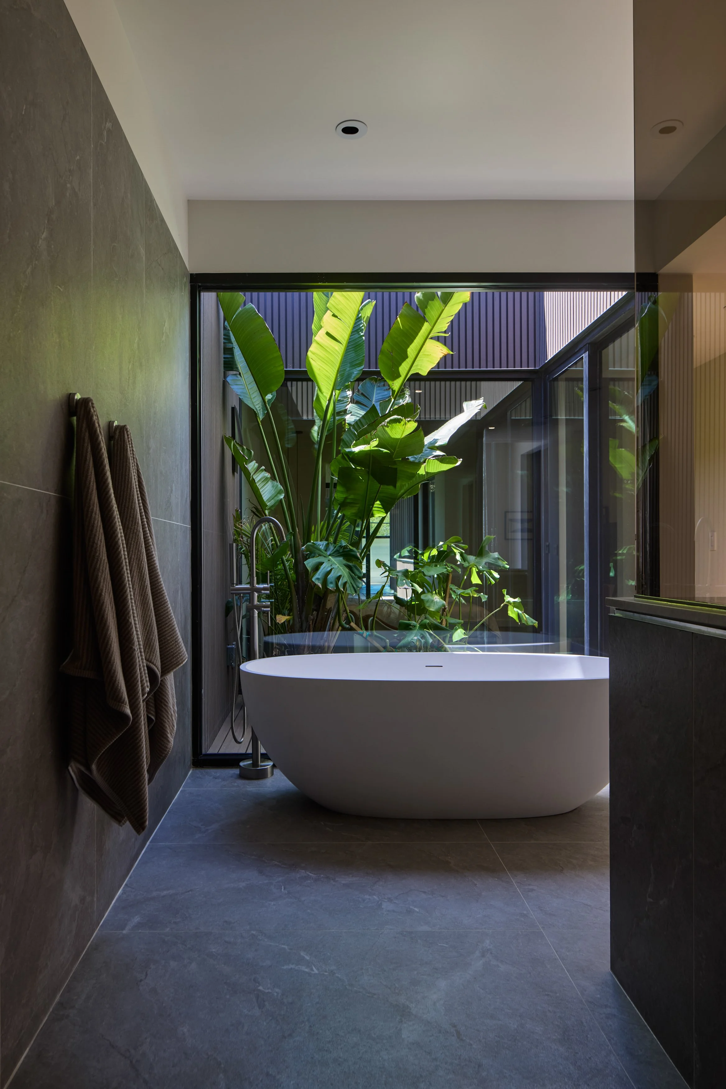 Modern bathroom with gray tiled flooring and walls, a freestanding white bathtub, large windows with greenery outside, and beige towels hanging on the wall.