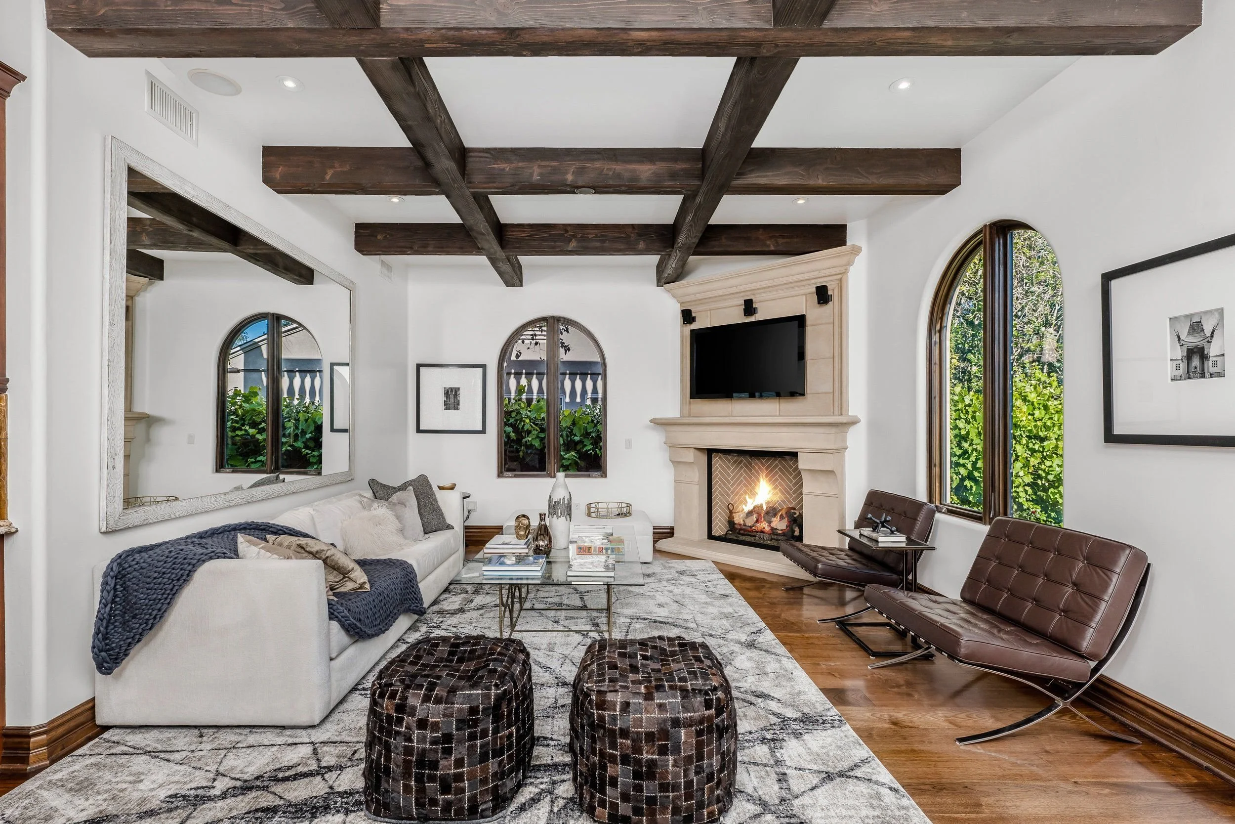 Living room with white sofa, dark wood ceiling beams, fireplace, large arched windows, brown leather chairs, decorative items on coffee table, and wall art.
