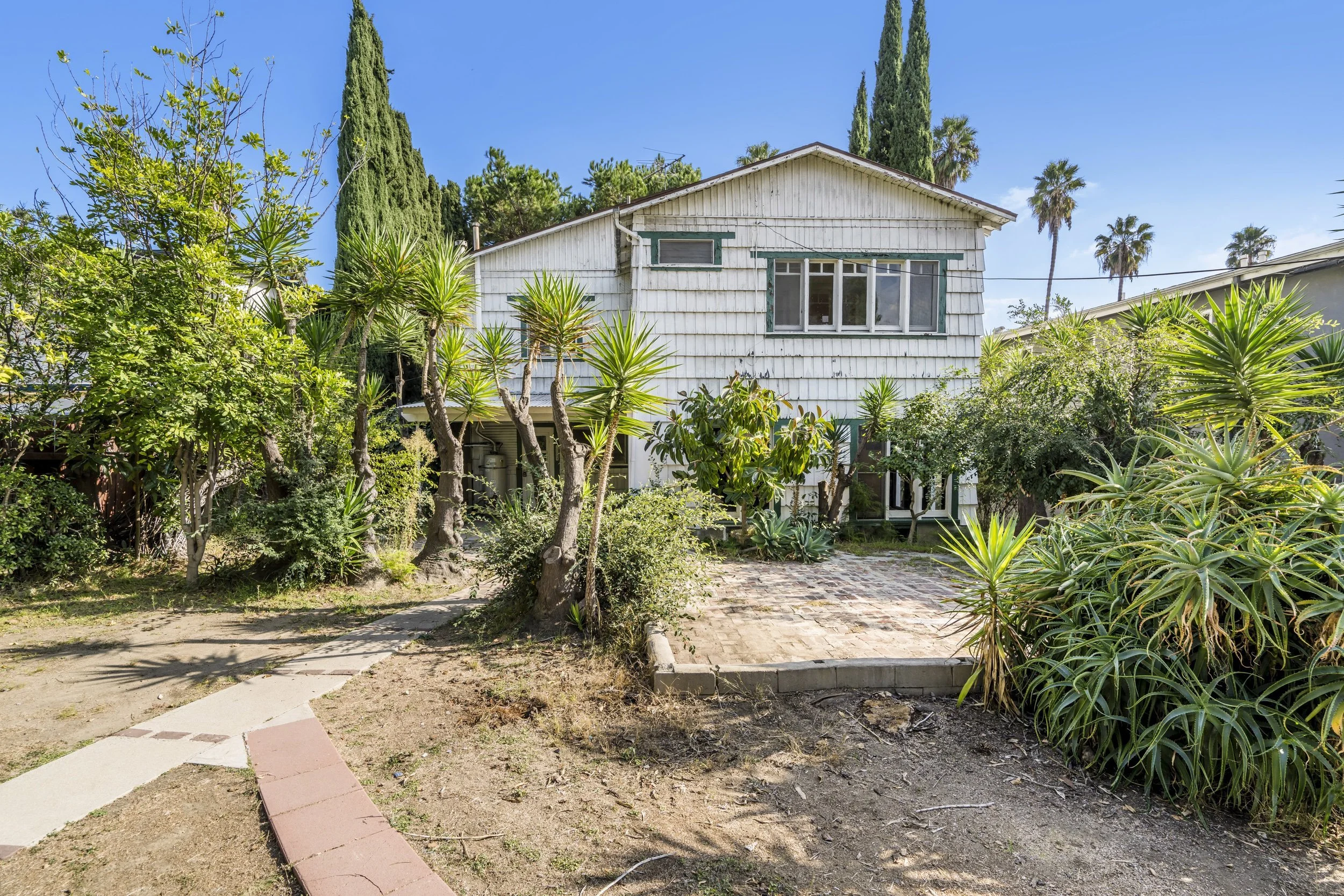 Front view of a two-story house with white wooden siding, surrounded by scattered desert plants and trees, under a clear blue sky.