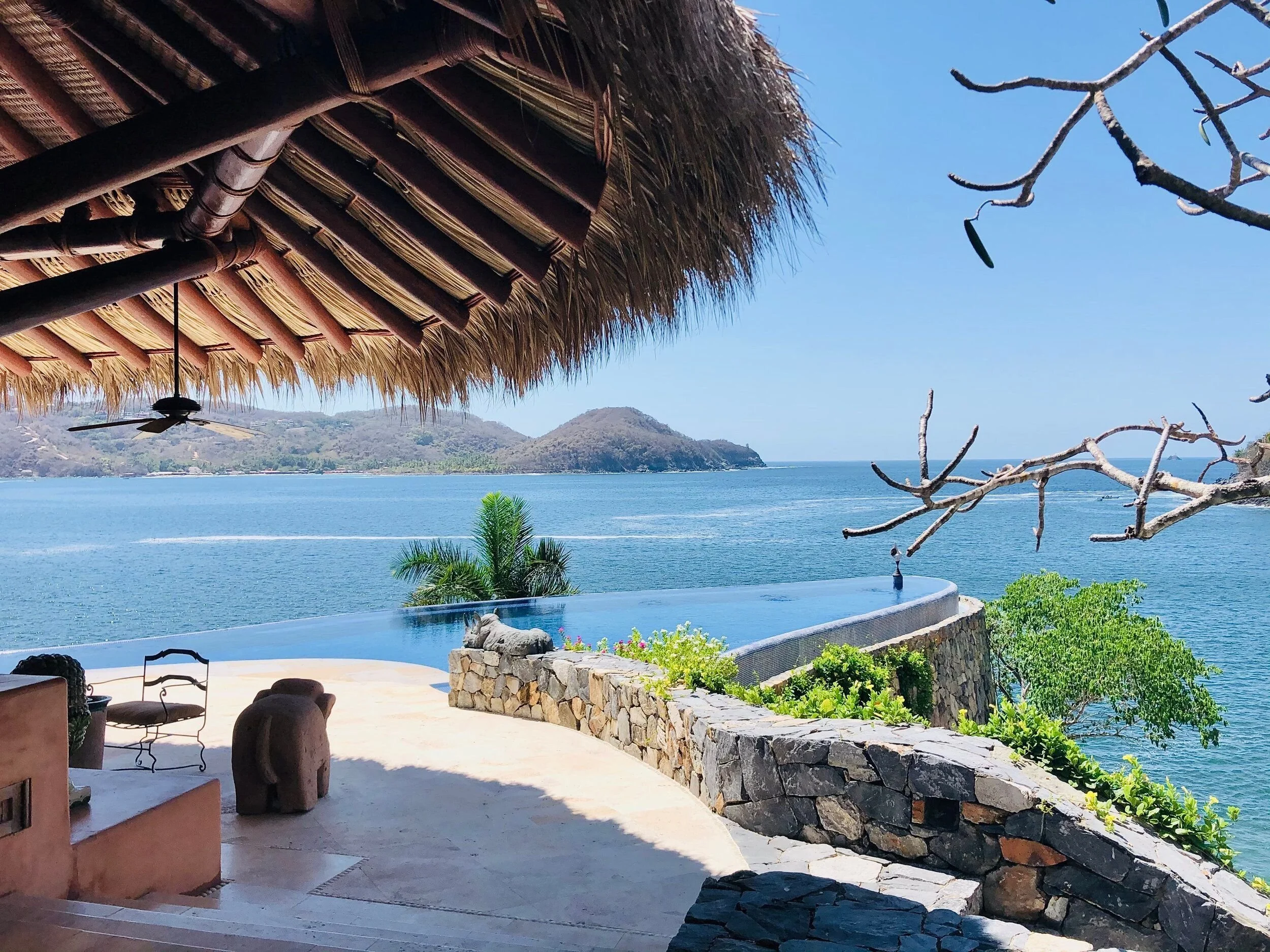 View from a shaded patio overlooking the ocean with mountains in the distance. The patio has wicker furniture, a stone wall, and a thatched roof overhead. A tree with bare branches extends into the frame.