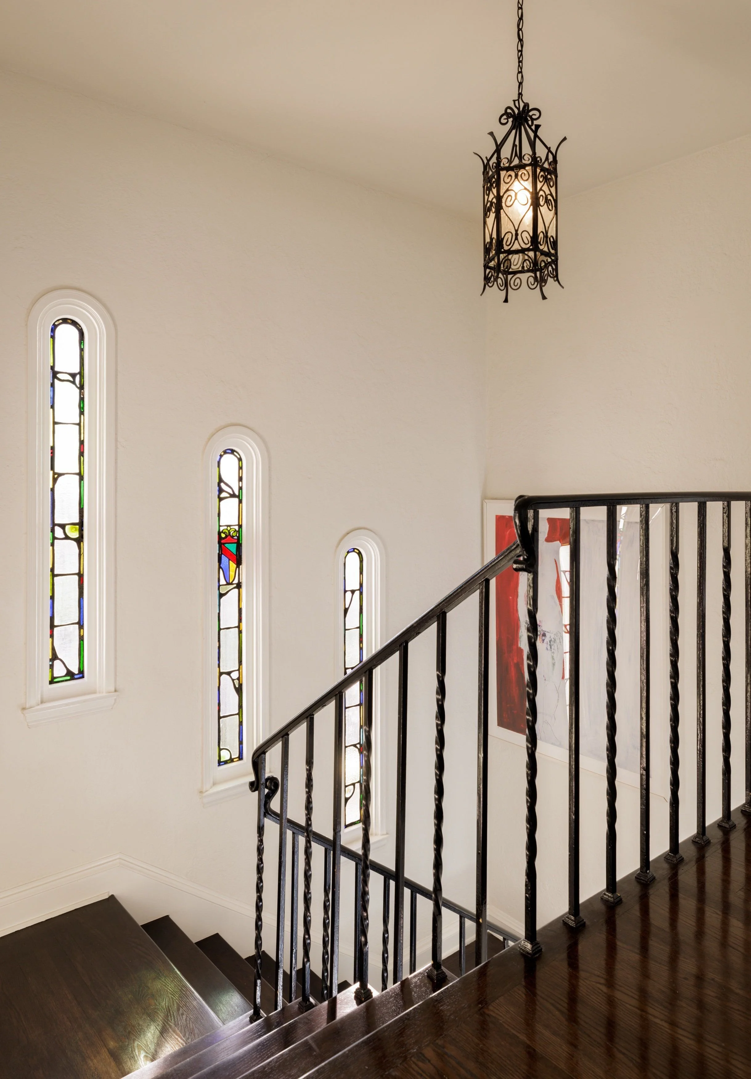 Interior view of a staircase with dark wood steps, a black wrought iron balustrade, stained glass windows on the wall, and a decorative lantern-style ceiling light.