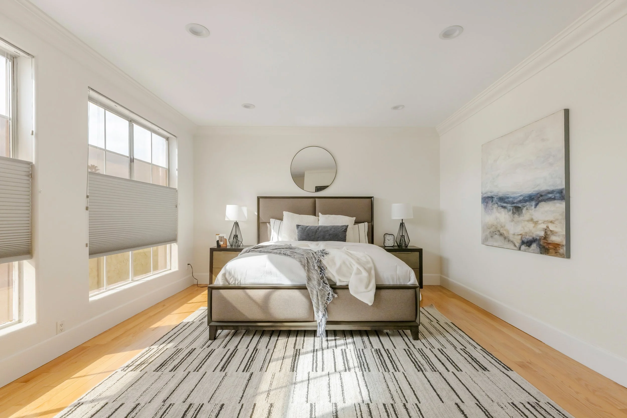 A minimalist bedroom with a bed, two nightstands with lamps, a round mirror above the headboard, large windows letting in natural light, a large painting on the wall, and striped area rug on wooden floor.