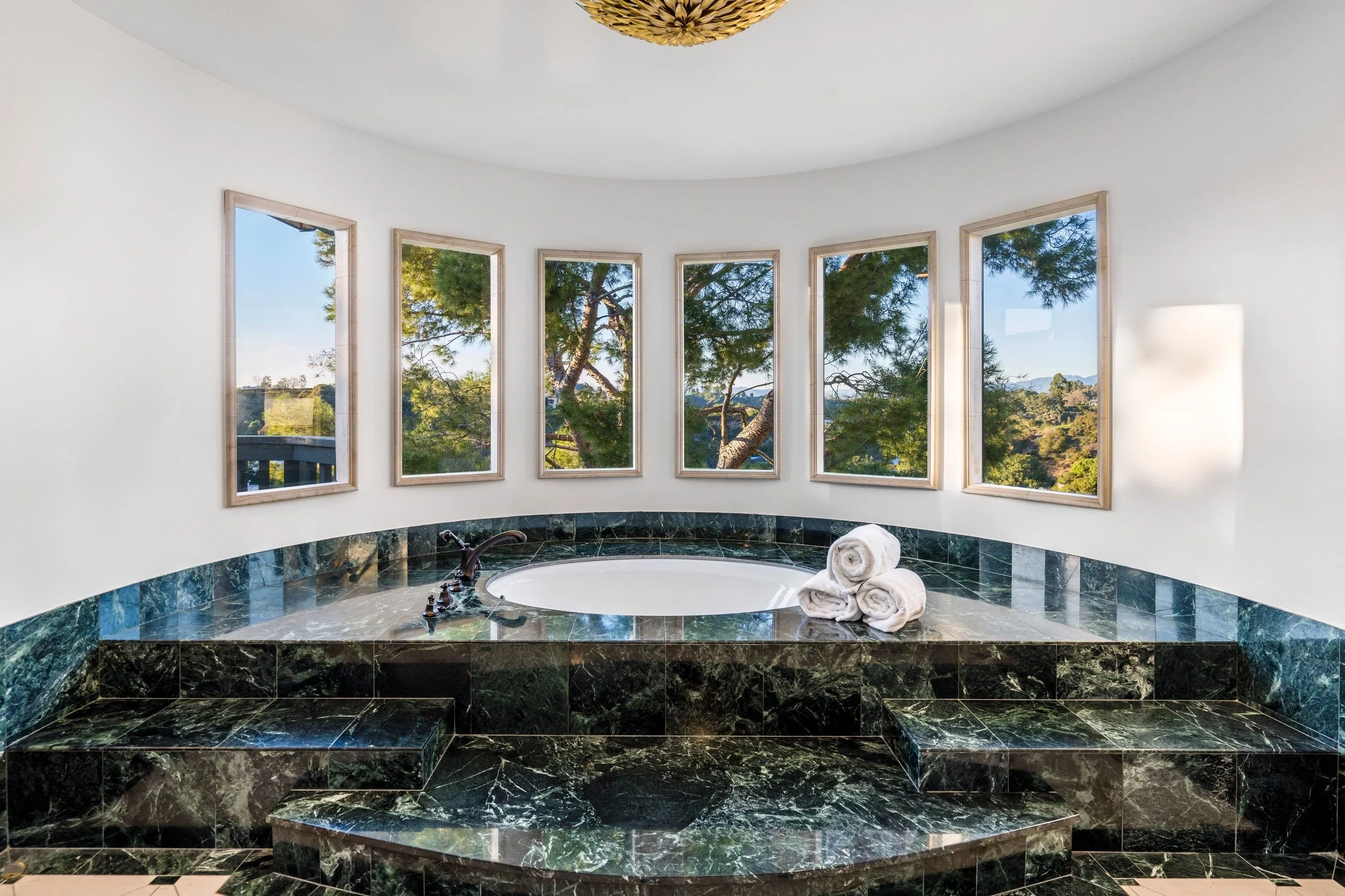 Round bathtub with dark green marble surround in front of a curved wall of windows showing trees and blue sky.