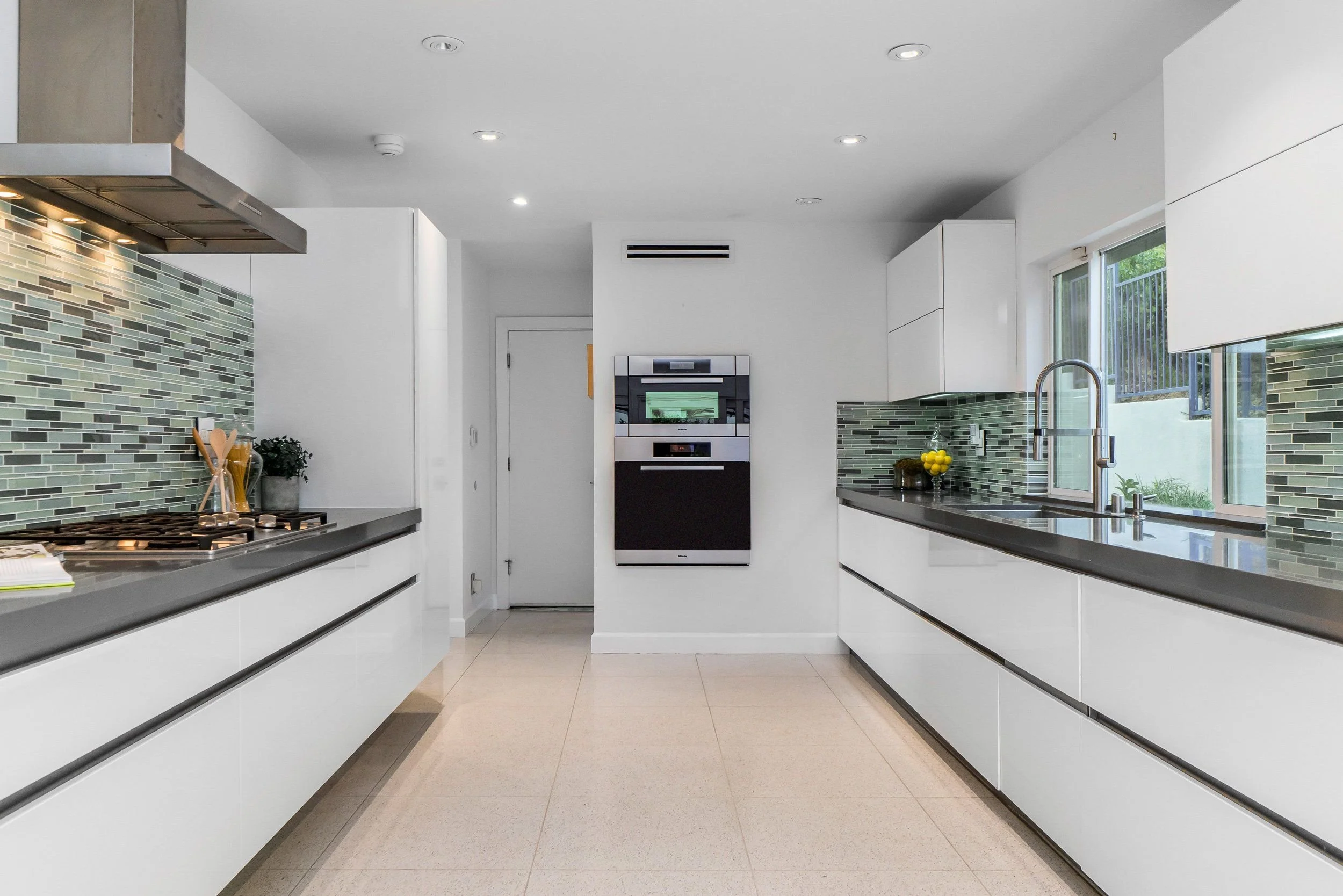 Modern kitchen with white cabinets, gray countertops, green glass tile backsplash, stainless steel appliances, a window above the sink, and beige tile floors.