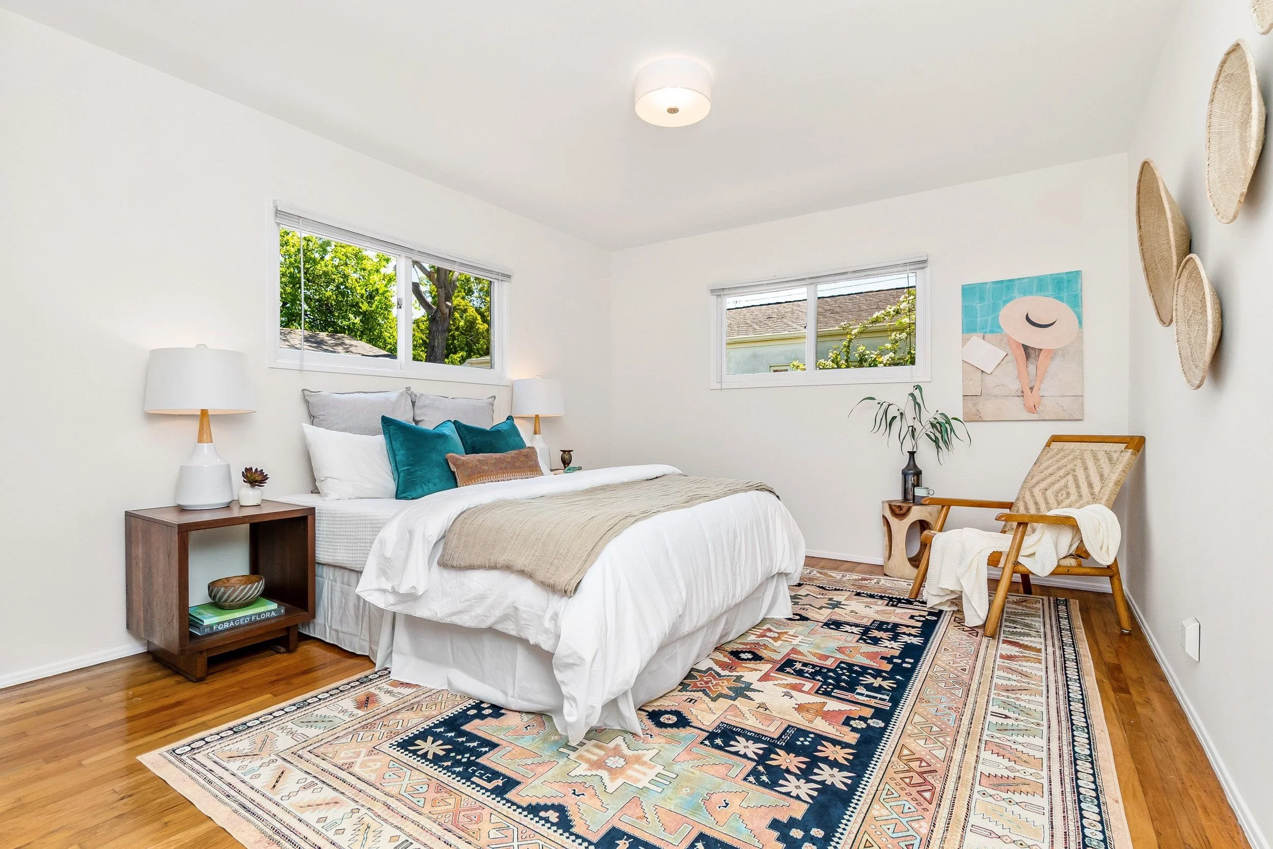 A bedroom with a white bed, blue and white pillows, a wooden bedside table with a lamp and books, and a colorful patterned rug. There are two windows showing green trees and a house roof outside. The wall has a painting and three woven wall baskets. 