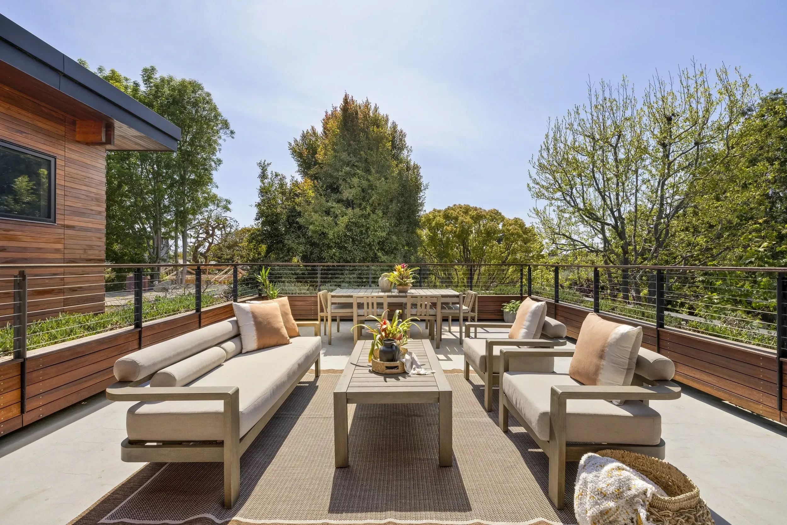 Outdoor patio with beige sofas and chairs, a wooden coffee table with plants, and a dining table, all bordered by a black railing and surrounded by green trees under a clear blue sky.