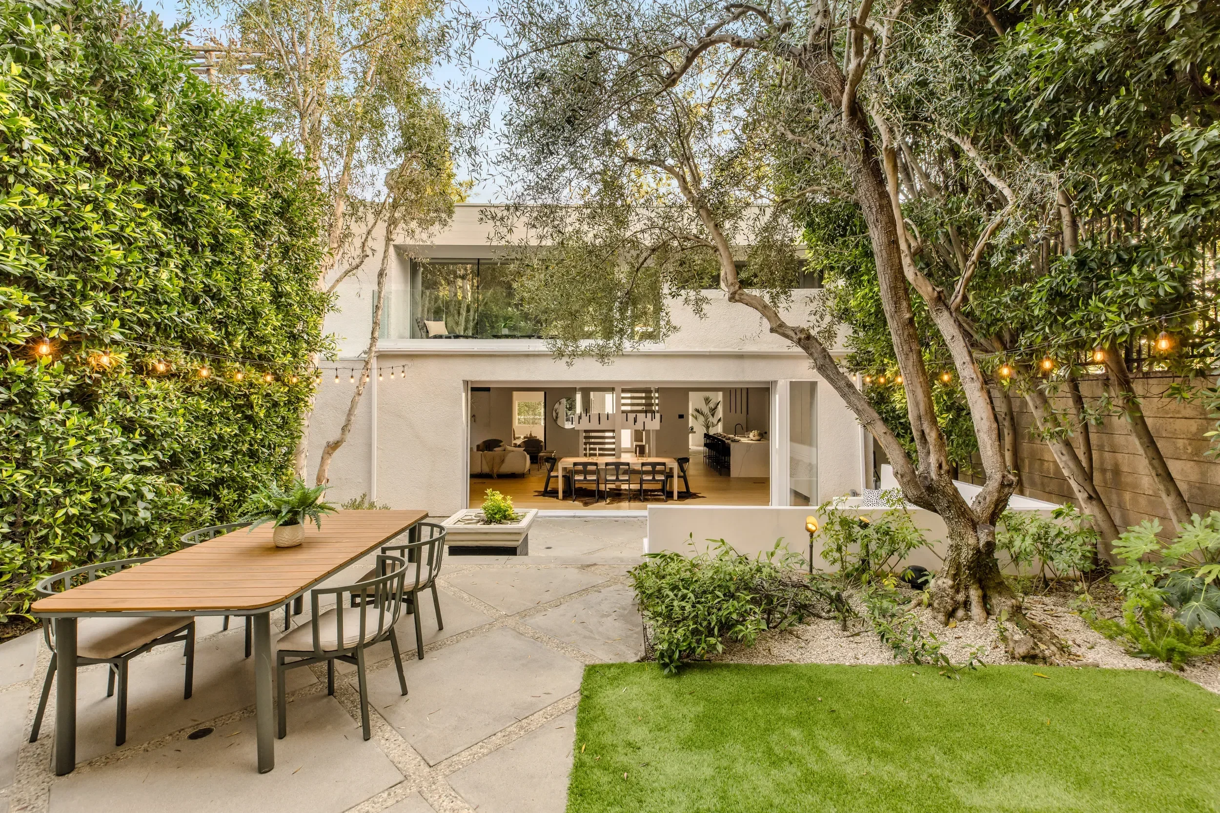 A modern backyard patio with a wooden dining table and black chairs, surrounded by lush greenery, trees, and string lights, with a view of a house's open sliding glass doors revealing an indoor living area.