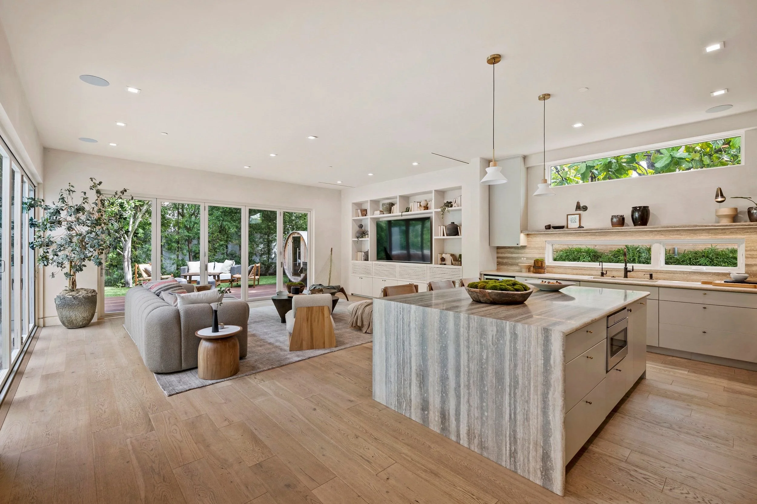 Open-concept living room and kitchen with large windows, white walls, wooden floors, a gray sofa, bookshelves with a TV, and a kitchen island with a marble countertop.