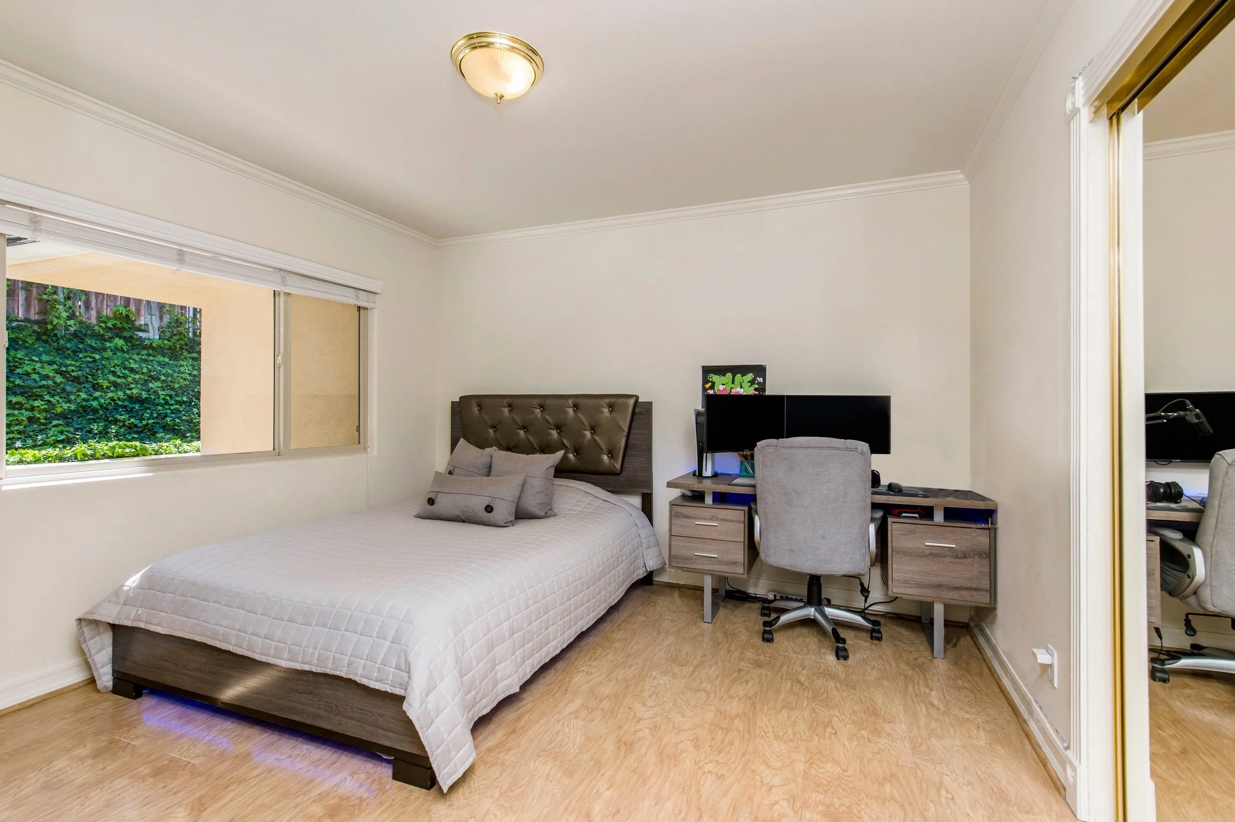 A bedroom with a bed, pillows, a window with green foliage outside, a workspace with a computer, a chair, and a closet with sliding door.
