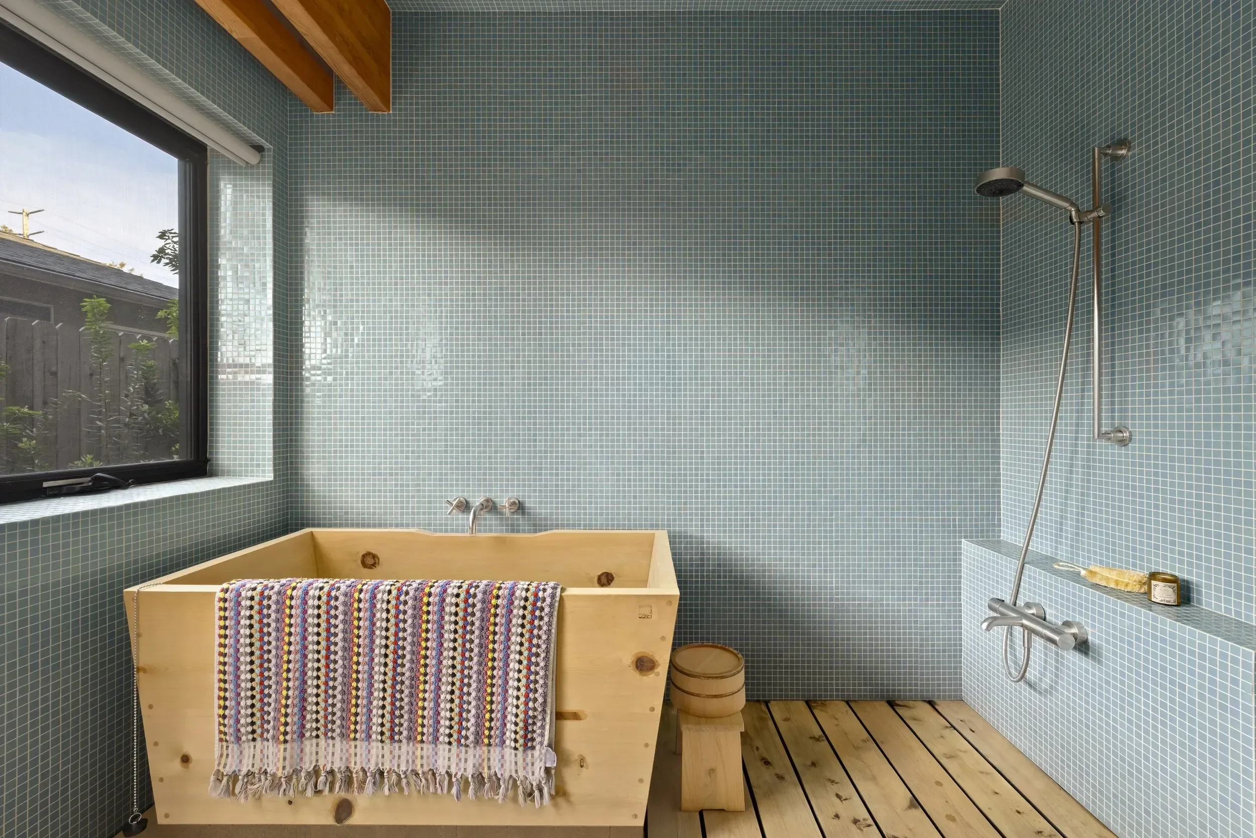 A bathroom with blue mosaic tile walls, a window, and a walk-in shower with a rain showerhead. There is a wooden bathtub with a colorful striped towel hanging on its side, and a small wooden stool with a candle and a bucket on the floor. The floor is