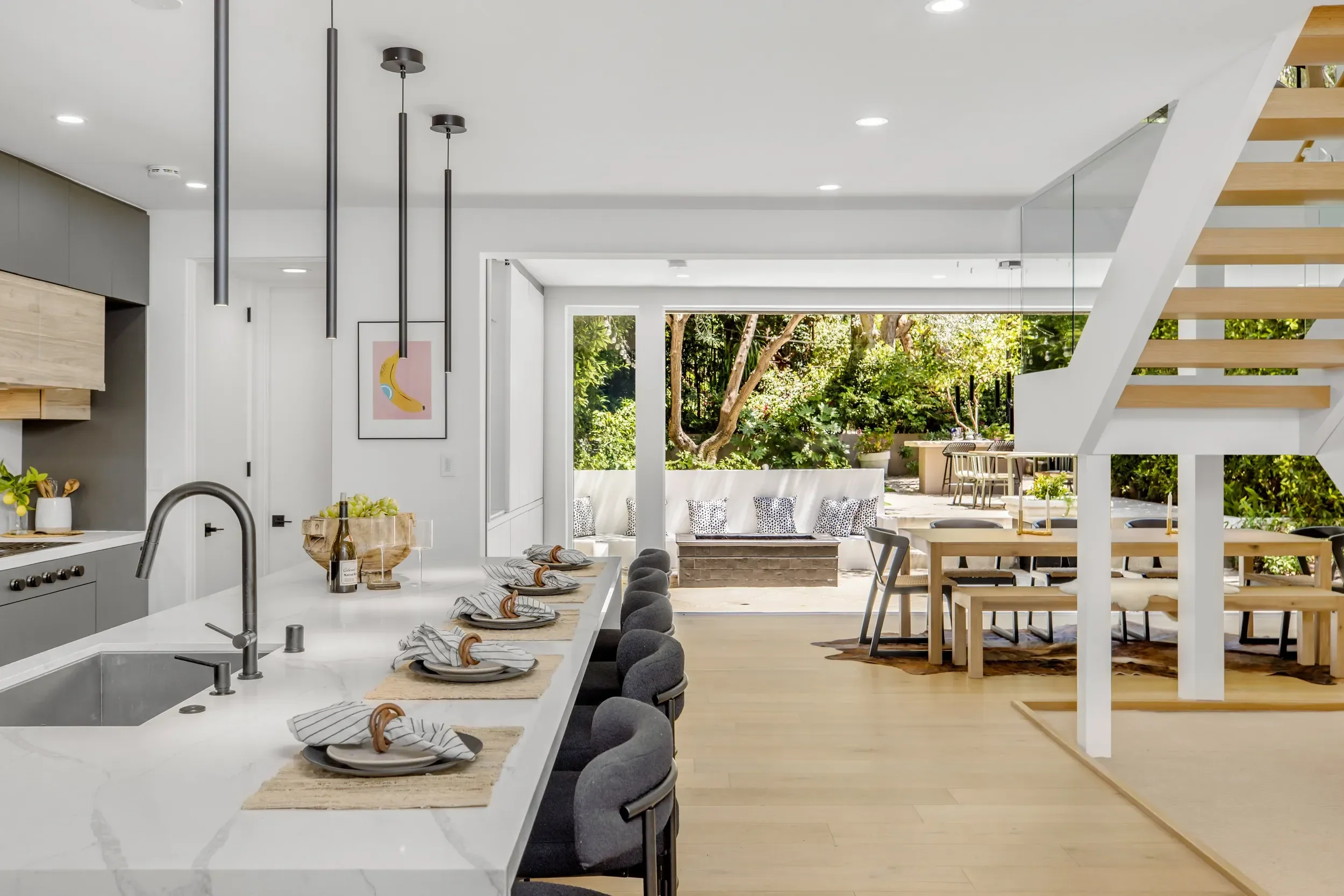 Modern kitchen and dining area with white countertops, gray bar stools, a large window, and a view of a lush backyard with trees and patio furniture.