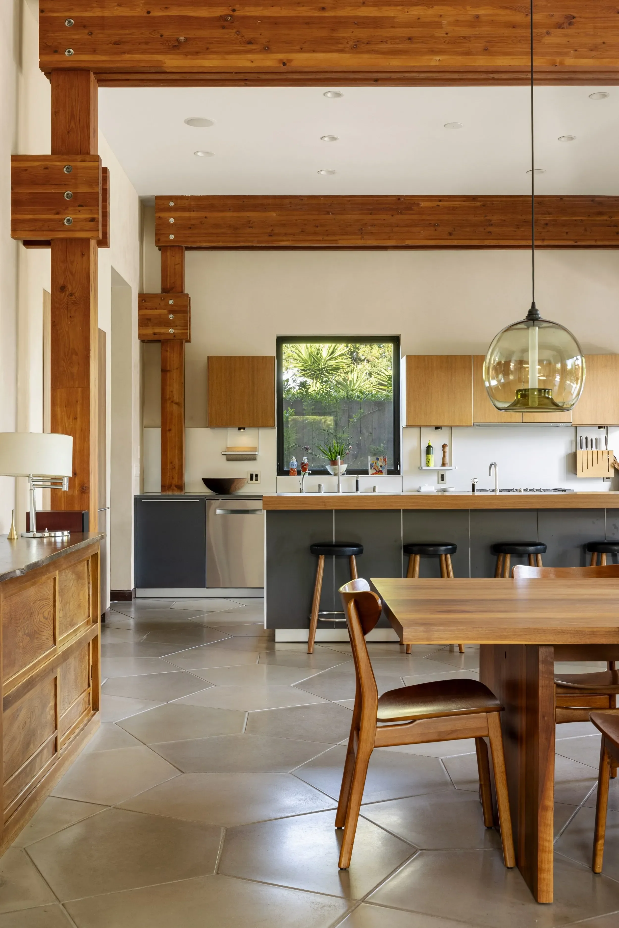 Modern kitchen with wooden beams, beige tiled floor, and wooden dining table with chairs. Kitchen cabinets are wood with a black countertop, and a large window shows greenery outside. A pendant light hangs over the table.