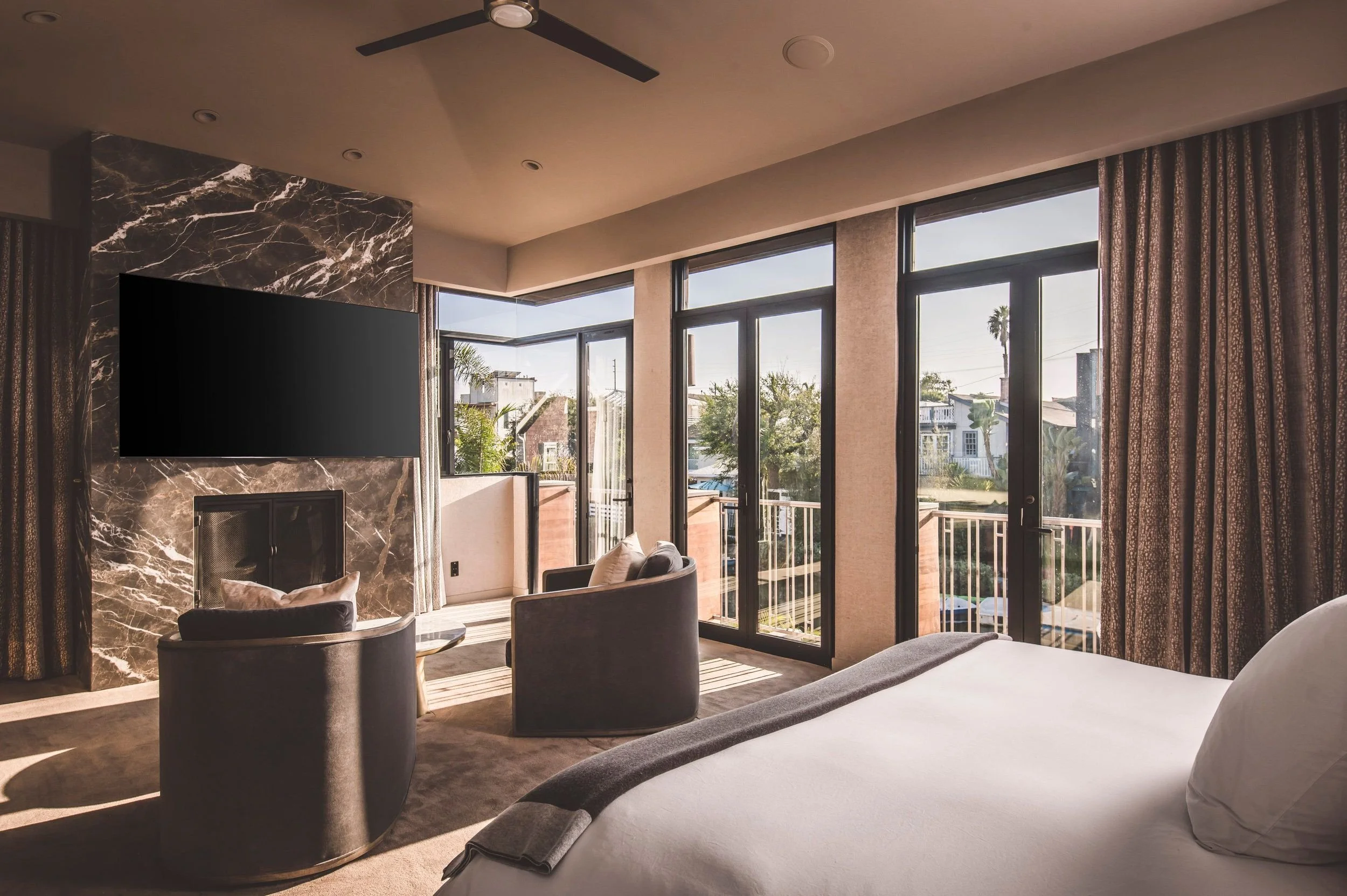 A modern living room with large floor-to-ceiling windows and sliding glass doors, revealing a balcony and neighborhood view. The room features a flat-screen TV mounted on a marble accent wall, a beige couch with pillows, a patterned curtain, and chai