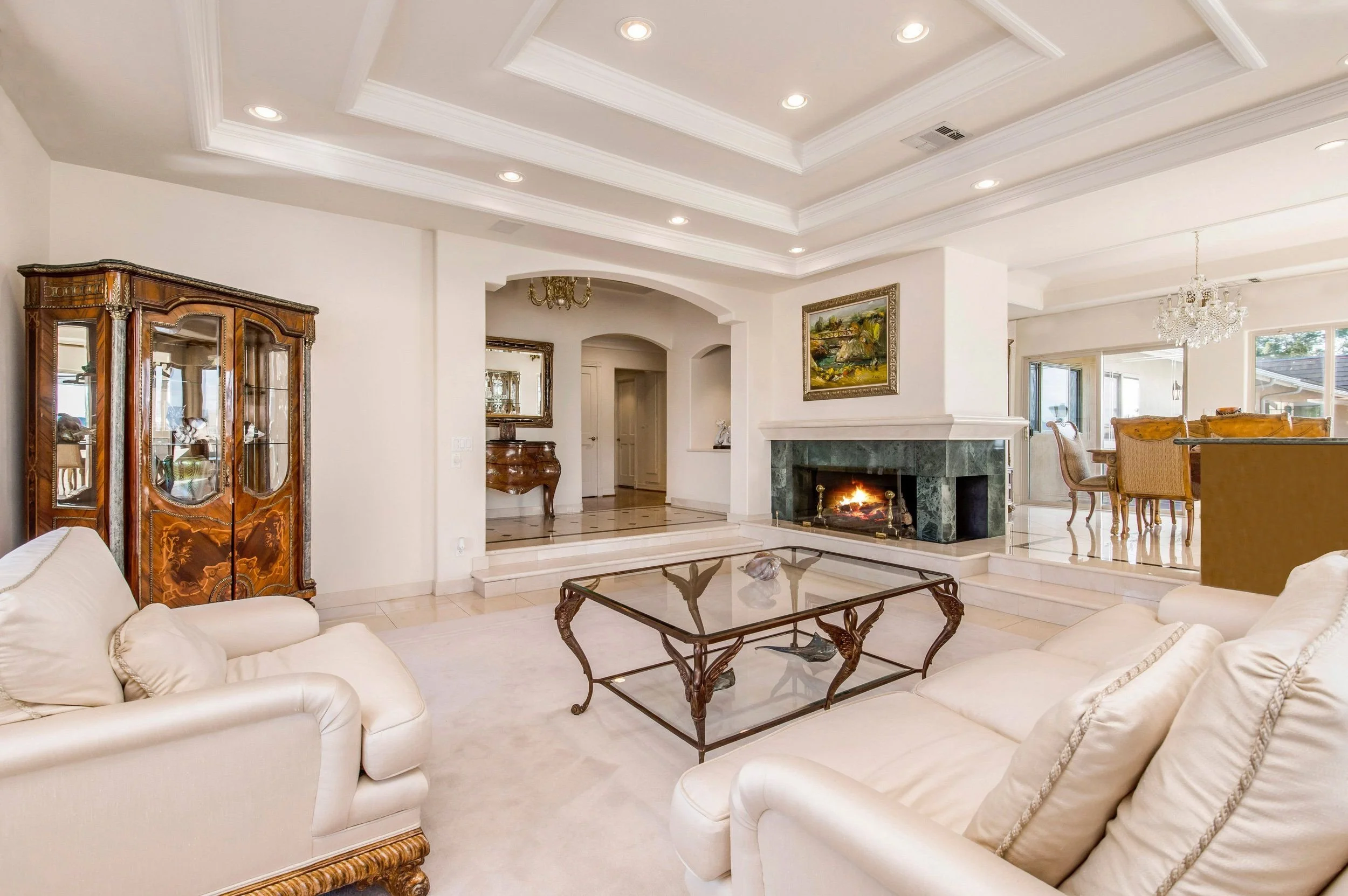 Living room with white couches, glass coffee table, fireplace, chandelier, and traditional wooden furniture.