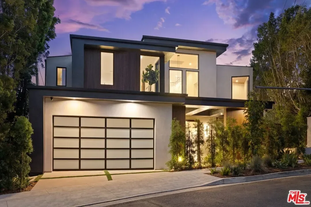 Modern two-story house with a white exterior, large windows, a garage with frosted glass panels, and landscaped front yard, illuminated at dusk.