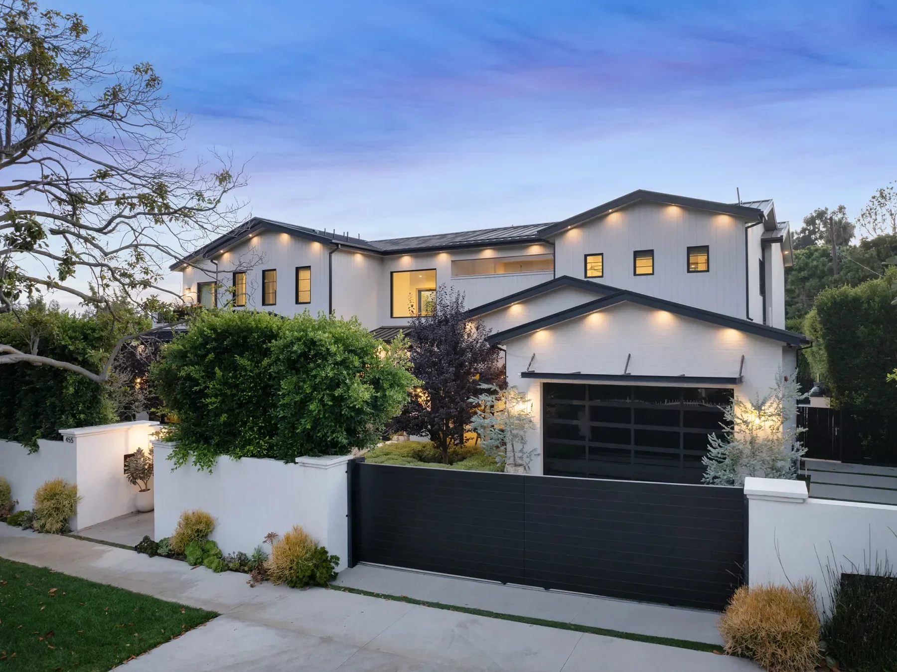 Modern two-story white house with black accents, surrounded by greenery and a white fence, during twilight with exterior lights on.