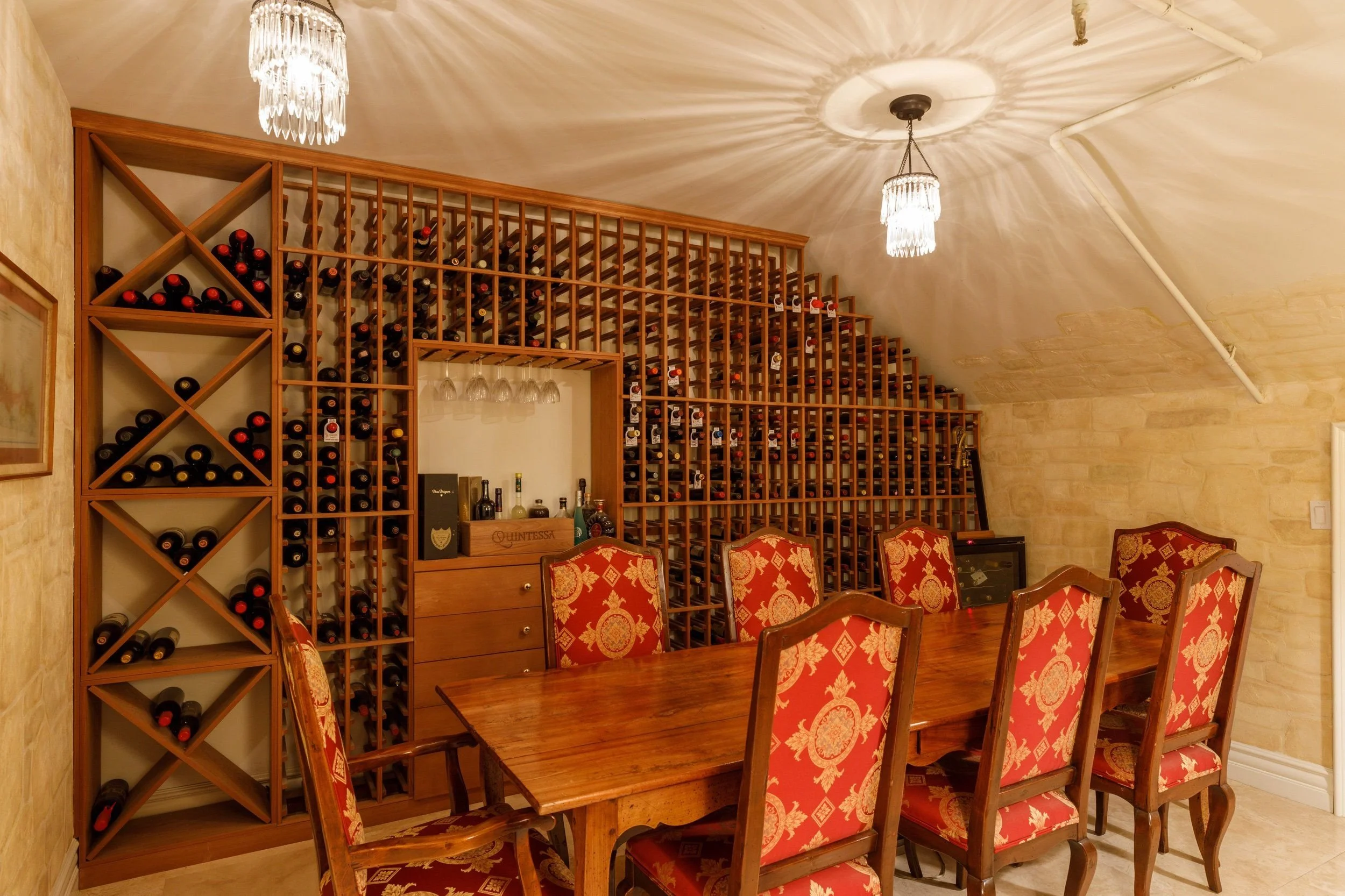 A wooden wine rack filled with wine bottles in a dining room with a large wooden table and chairs with red and gold upholstery, a chandelier, and a light-colored stone wall.