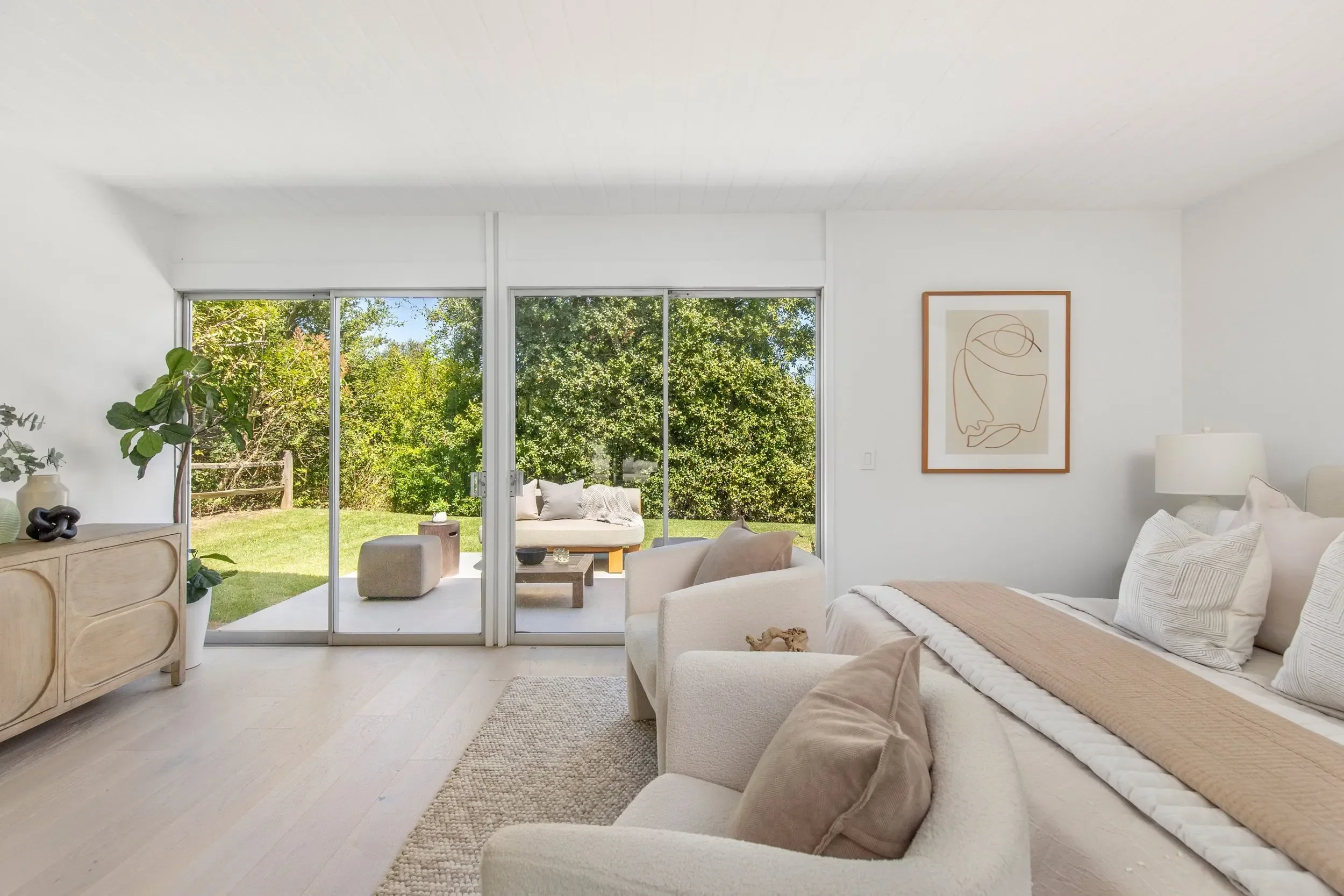 Living room with white walls and furniture, large sliding glass door leading to a backyard with trees, framed abstract art on the wall, and decorative plants and objects.