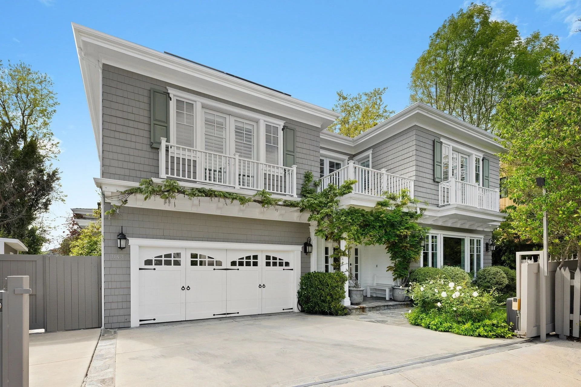 Two-story modern house with gray siding, white trim, and a two-car garage, surrounded by greenery and trees, under a clear blue sky.