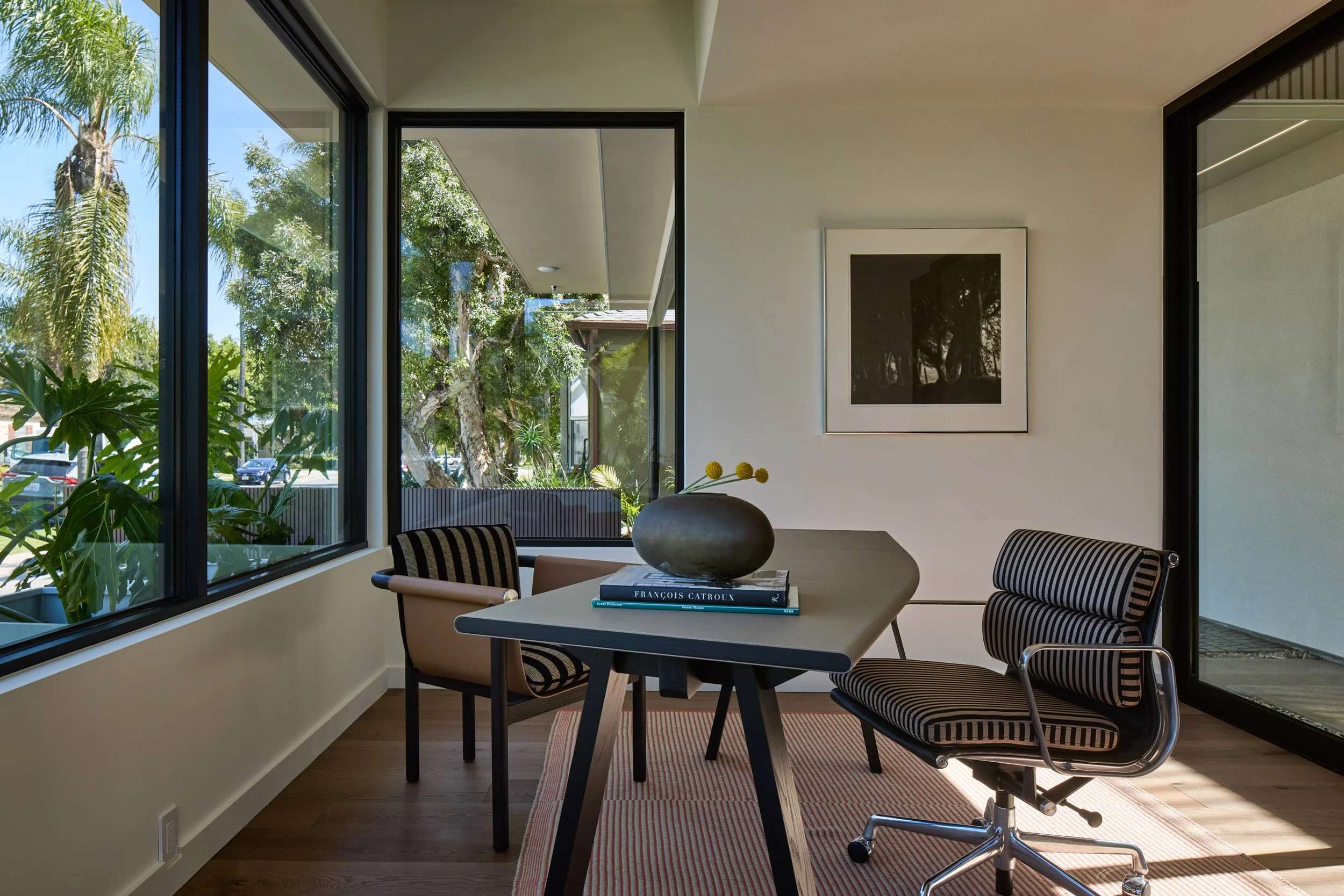 Modern dining area with a round table, four chairs, a large vase with flowers, and large windows showing greenery outside.