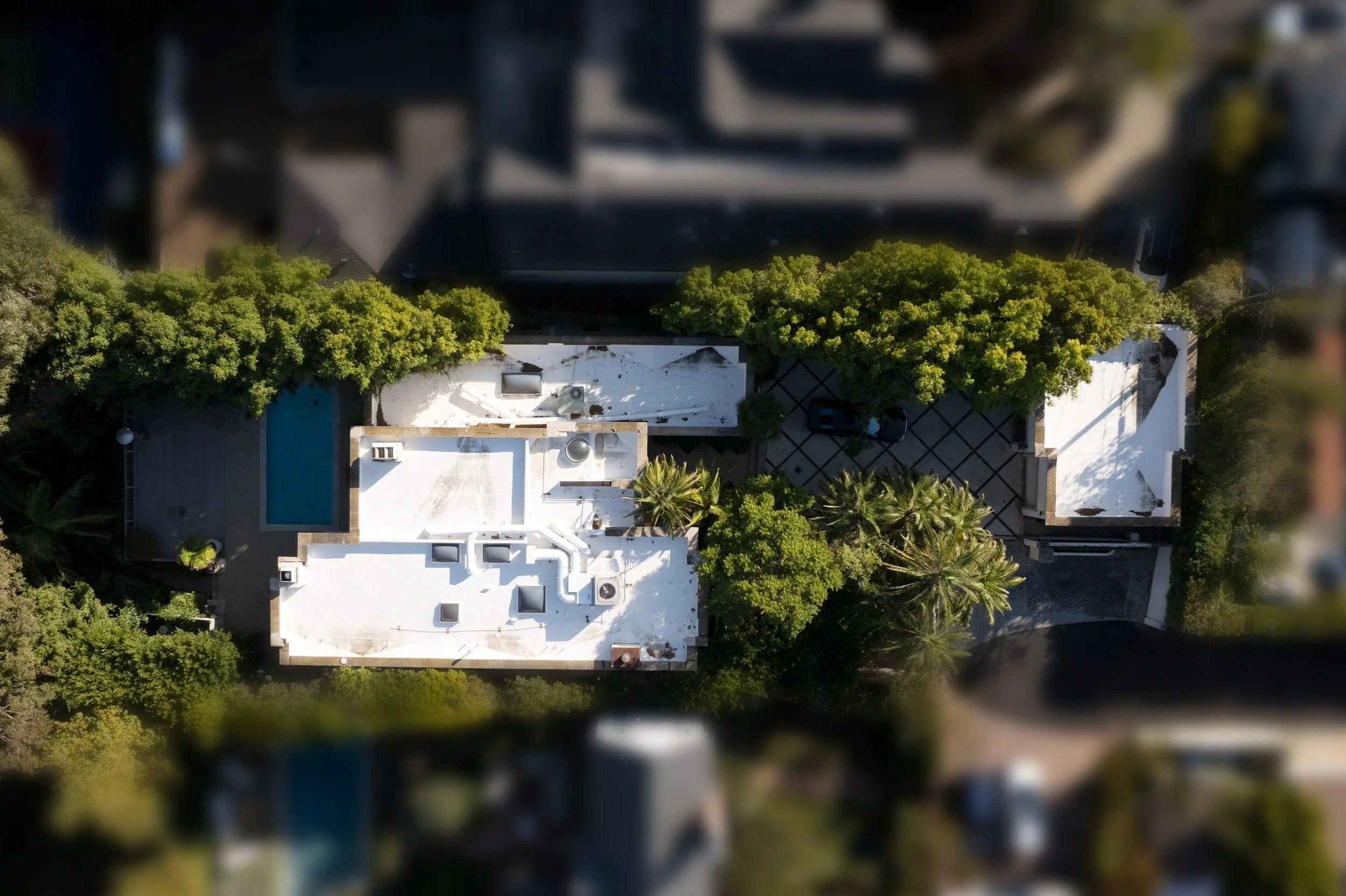 An aerial view of a modern white building with a flat roof surrounded by green trees, a swimming pool, and a parking area.