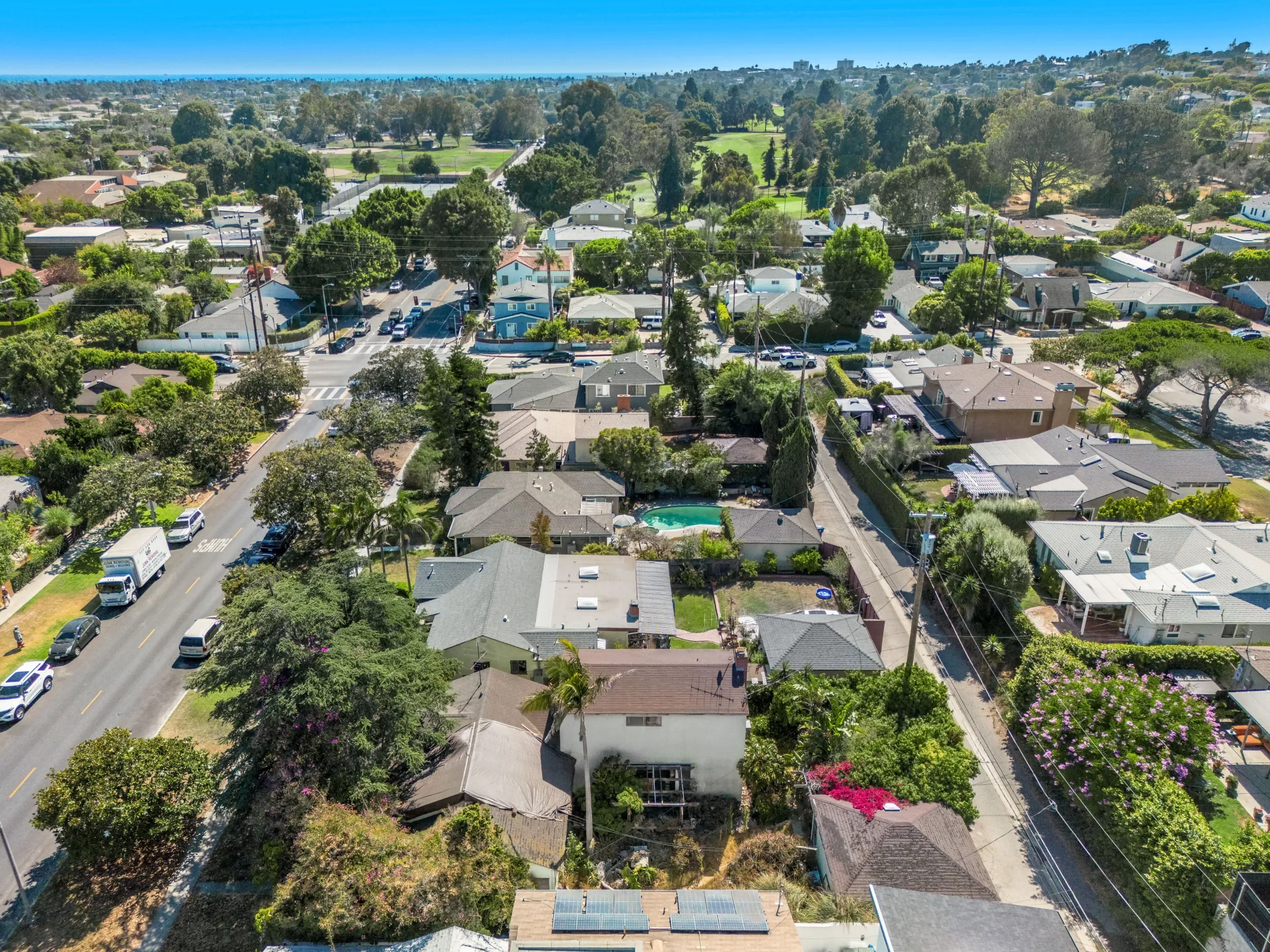 Aerial view of a suburban neighborhood with houses, a street, and a distant park with trees, under a clear blue sky.
