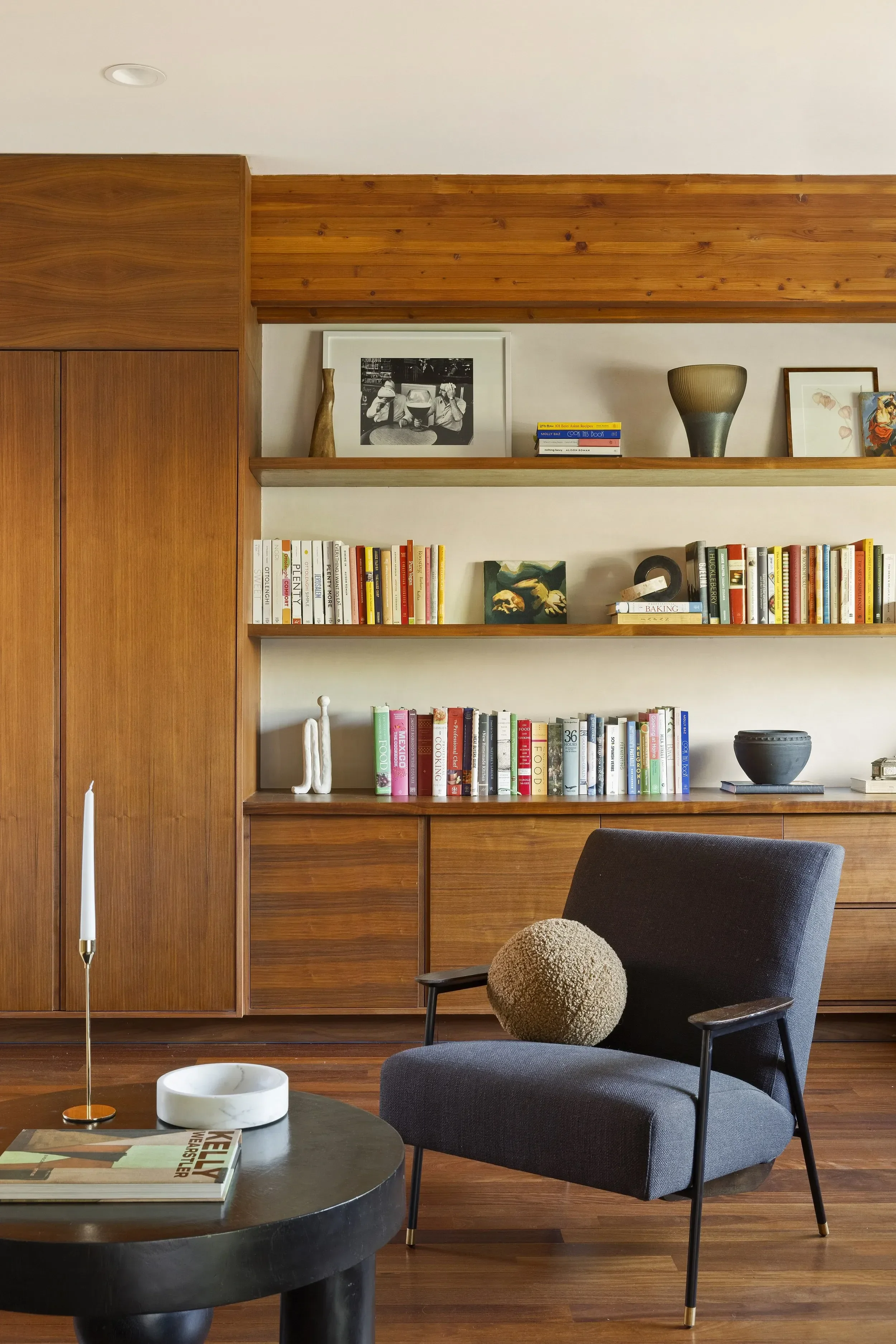 Living room with wooden cabinets, bookshelves filled with books, a black armchair with a round pillow, a black coffee table with a magazine, a white marble bowl, and a white candle. Decorative items and framed pictures on the shelves, a wooden ceilin