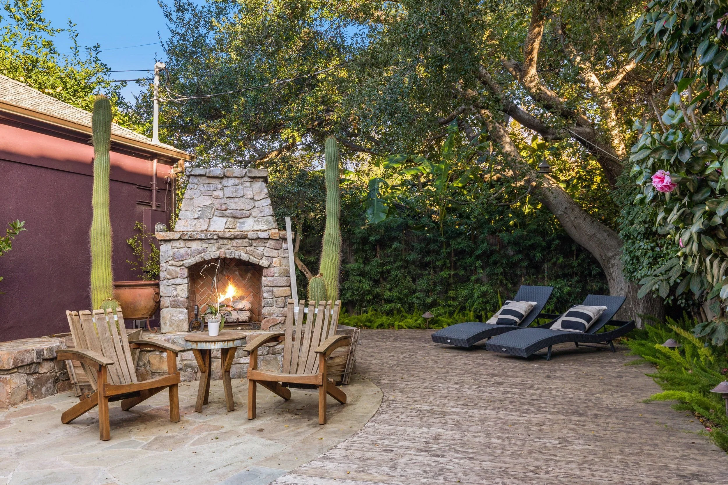 Backyard patio with a stone fireplace, potted plants, wooden chairs around a table, and lounge chairs on a brick patio surrounded by trees and greenery.