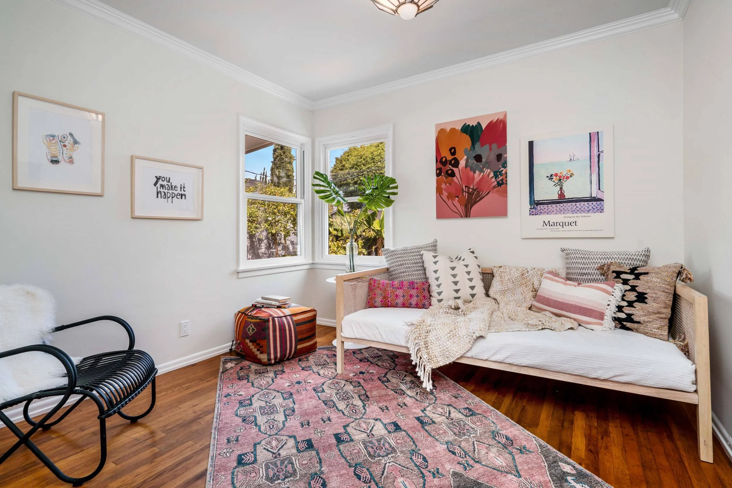 A cozy living room corner with a white couch covered with patterned pillows and a blanket, a colorful vintage-style rug, black metal chairs, a striped ottoman, a green plant in a glass vase on a side table, and framed artwork on the white walls near 