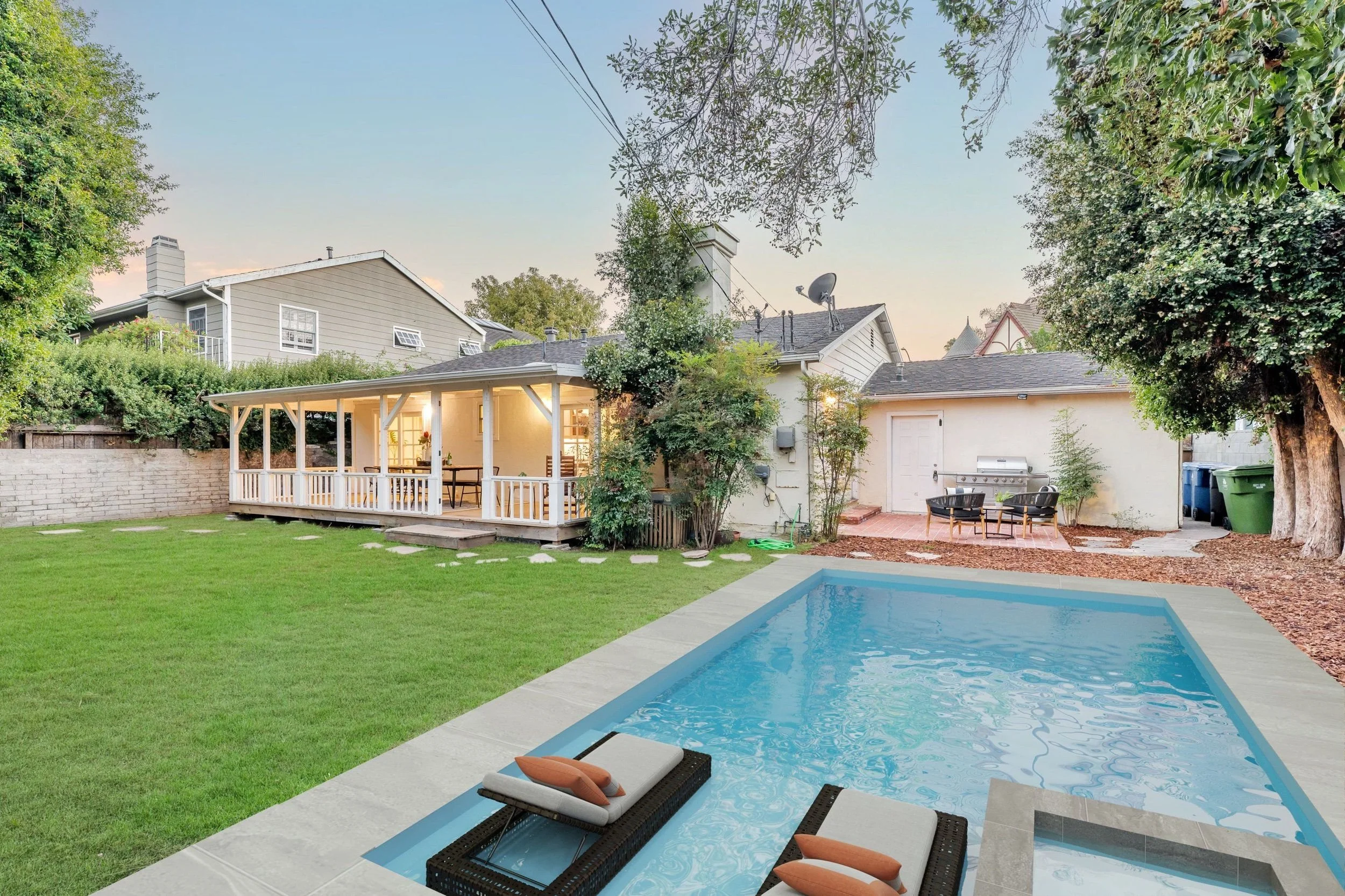 Backyard view with a swimming pool, lounge chairs, a covered patio, green grass, trees, and neighboring houses in the background during the evening.