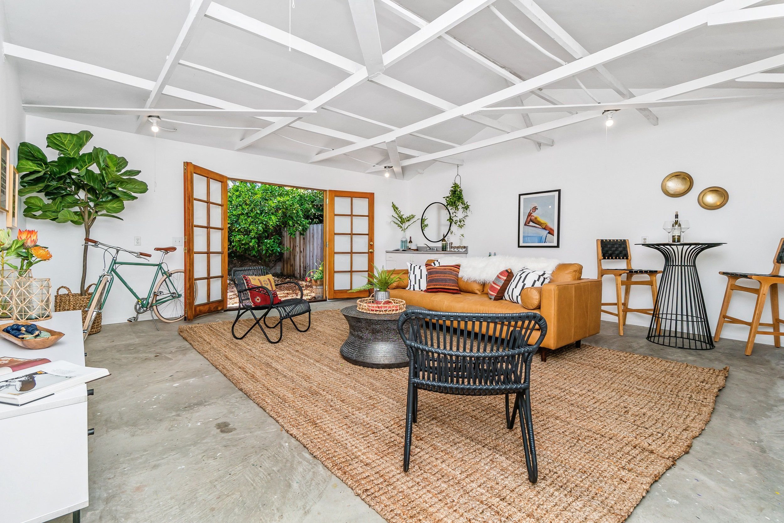 Living room with white walls and ceiling, open door leading to outdoor garden, tan sofa with patterned cushions, black armchairs, round coffee table, dining table with chairs, decorative plants, artwork on wall, and bicycle.