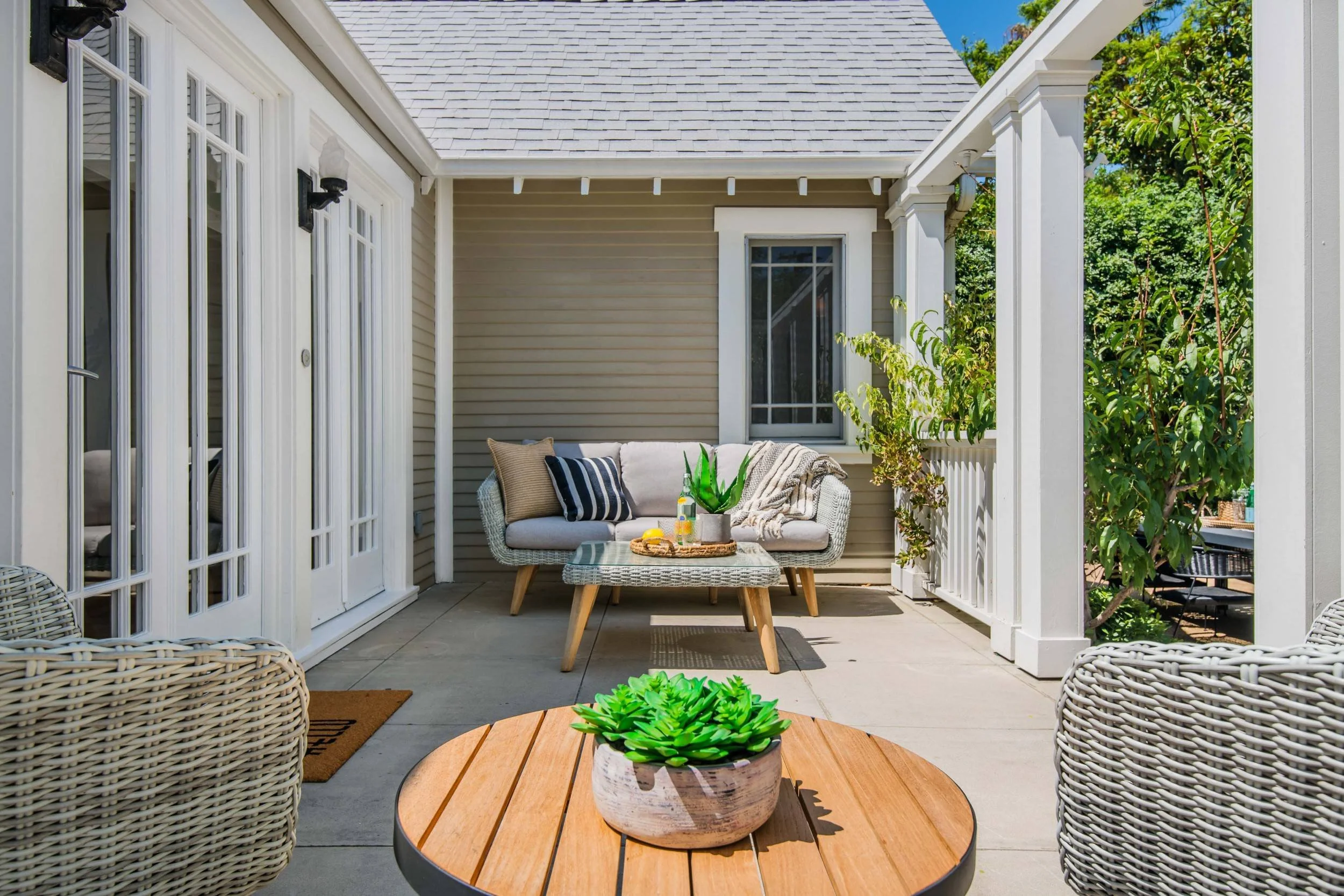 A cozy outdoor patio with a wicker sofa, a small wooden table with a potted succulent, and additional wicker chairs. The area is enclosed by a house with beige siding, white trim, and large glass doors. There's greenery in the background and a bright