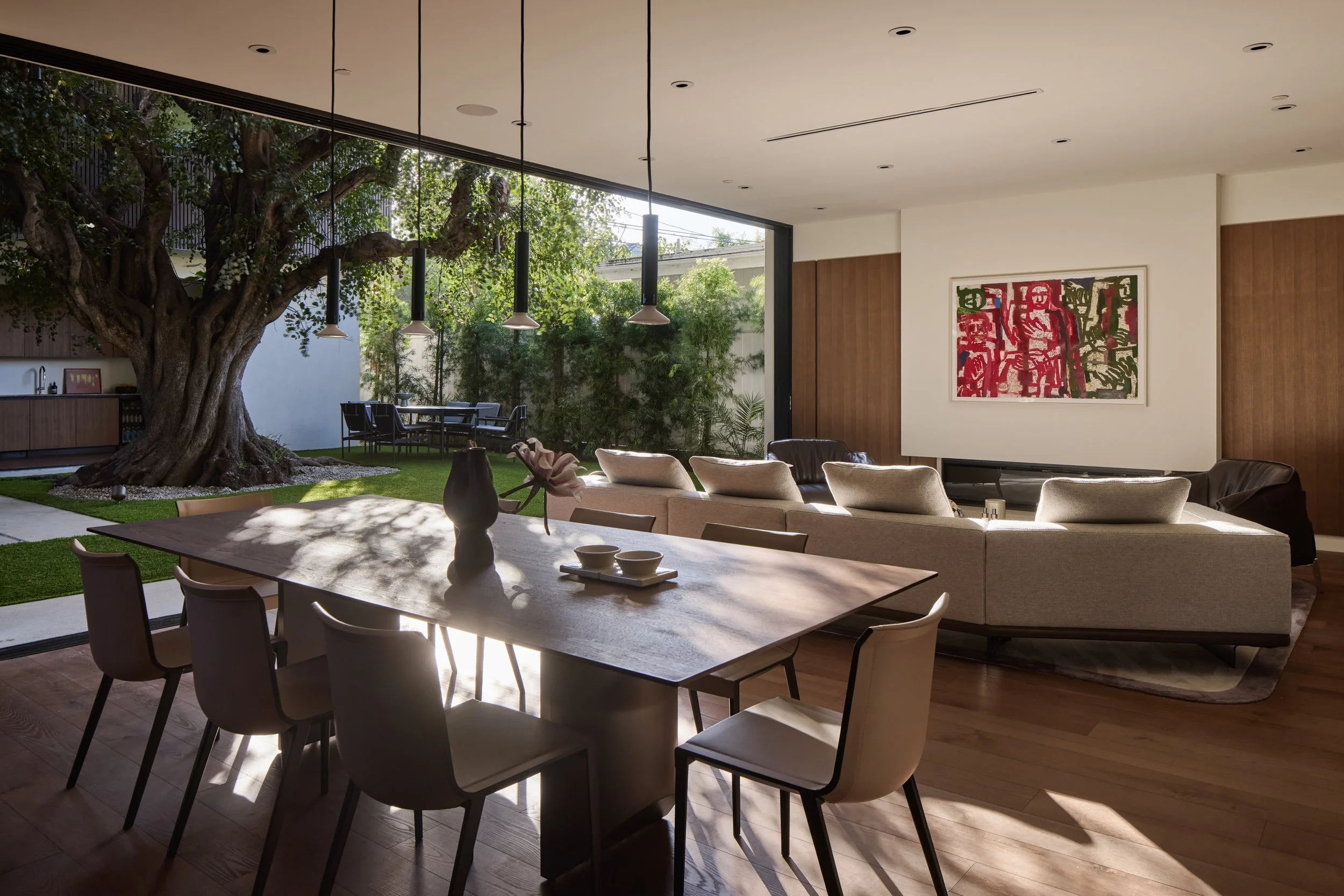 Modern living room with large open windows, a tree outside, and a dining table with chairs. There is a white couch with pillows, a wall with colorful abstract artwork, and pendant lights hanging from the ceiling.