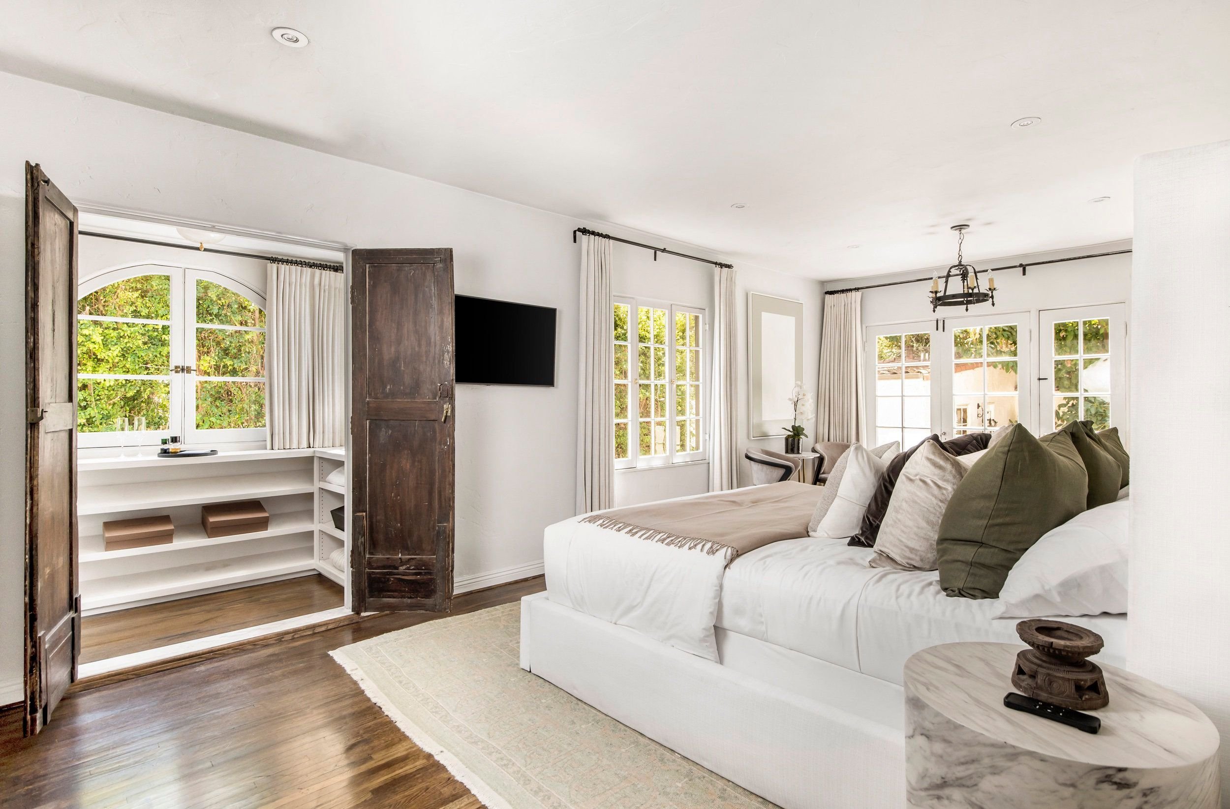 Bright living room with white walls, large windows with white curtains, a white sofa with multiple pillows, a wooden side table with decorative items, a wall-mounted television, and a chandelier hanging from the ceiling.