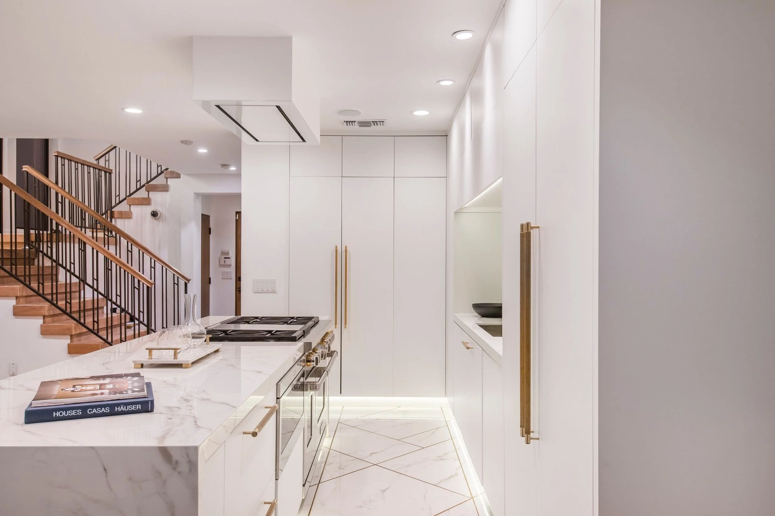 Modern kitchen interior with white cabinetry, marble countertops, a gas stove, and wooden stairs with black metal railing in the background.