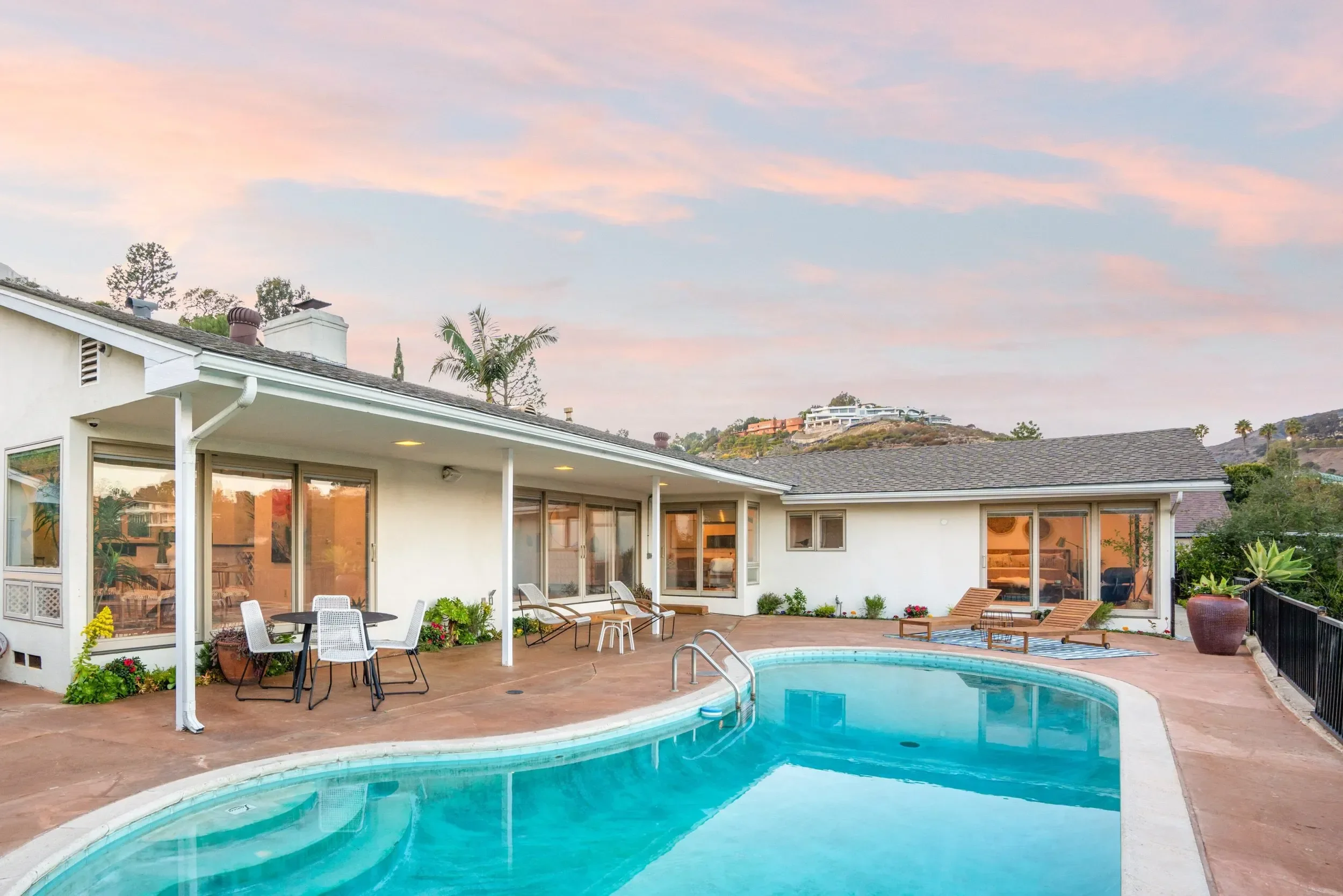The image shows a backyard with a swimming pool, patio furniture including dining table and chairs, lounge chairs, and lush greenery, with a view of houses and hills in the background during sunset.