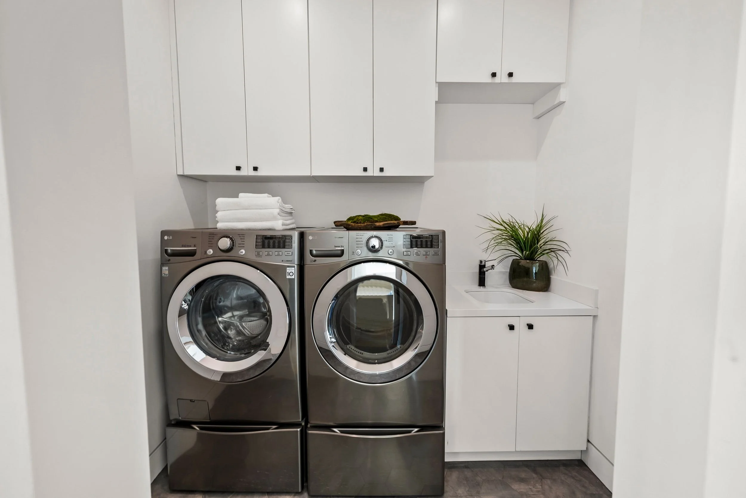 A laundry room with a stacked washer and dryer, white cabinetry, a small sink, a potted plant, and neatly folded towels on top of the washer.