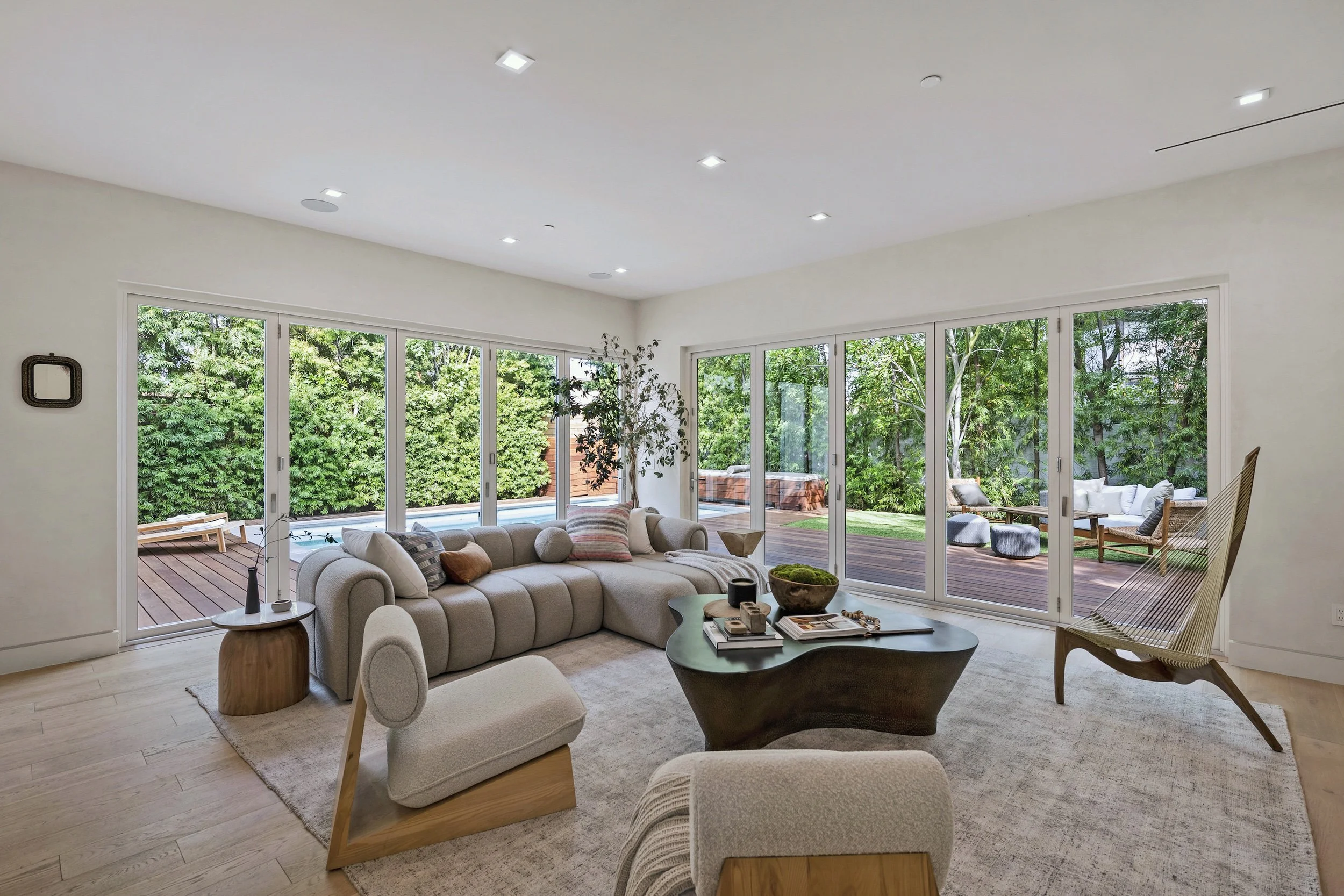 Living room with large glass doors overlooking a backyard with trees and patio furniture, beige sofa with pillows, wooden side table, coffee table, and modern chairs.
