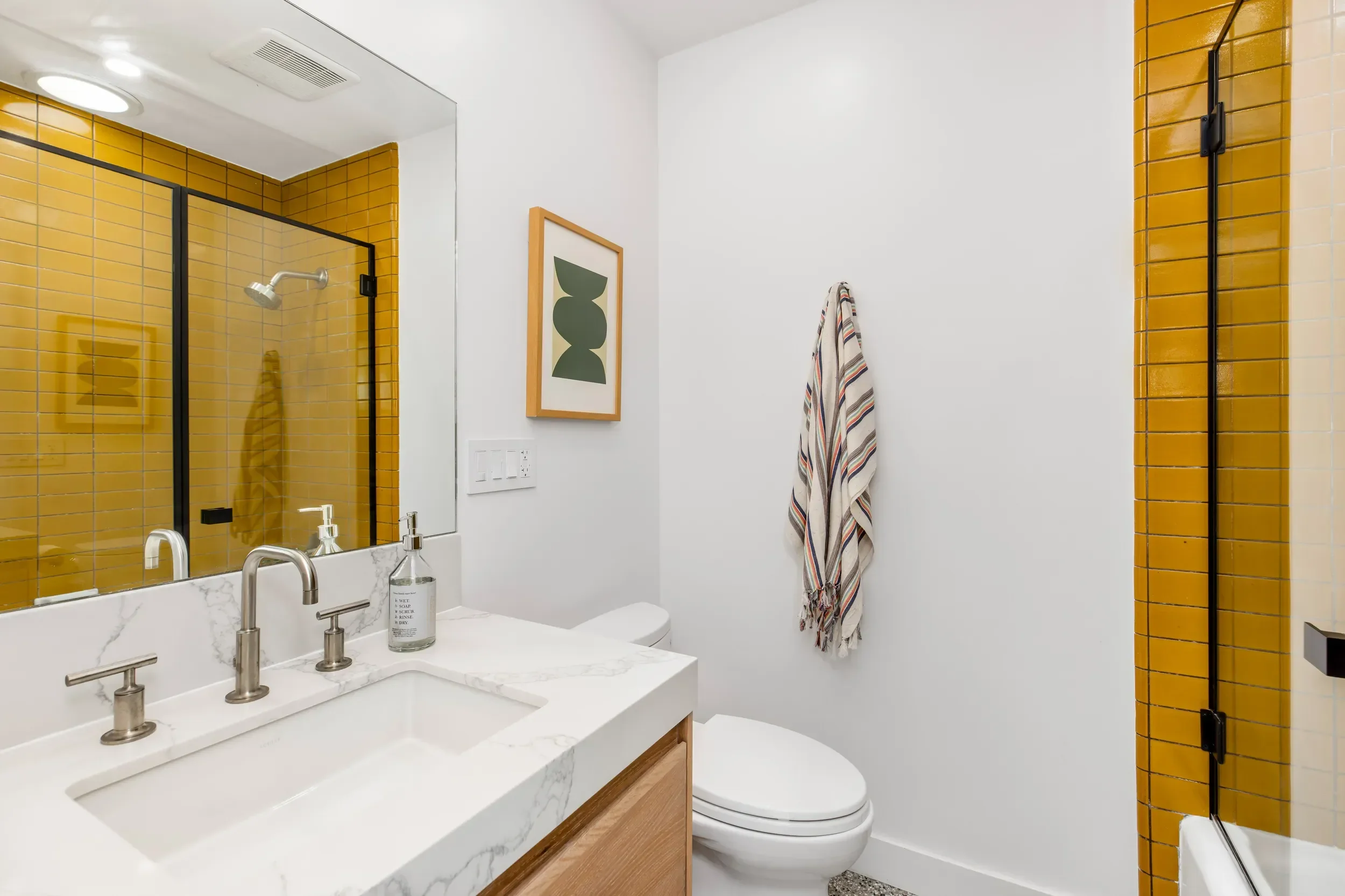 A modern bathroom with a yellow tiled shower stall, a white vanity with a marble countertop, a rectangular mirror, and a white toilet. A striped towel hangs on the wall, and a framed abstract art piece is above the toilet.